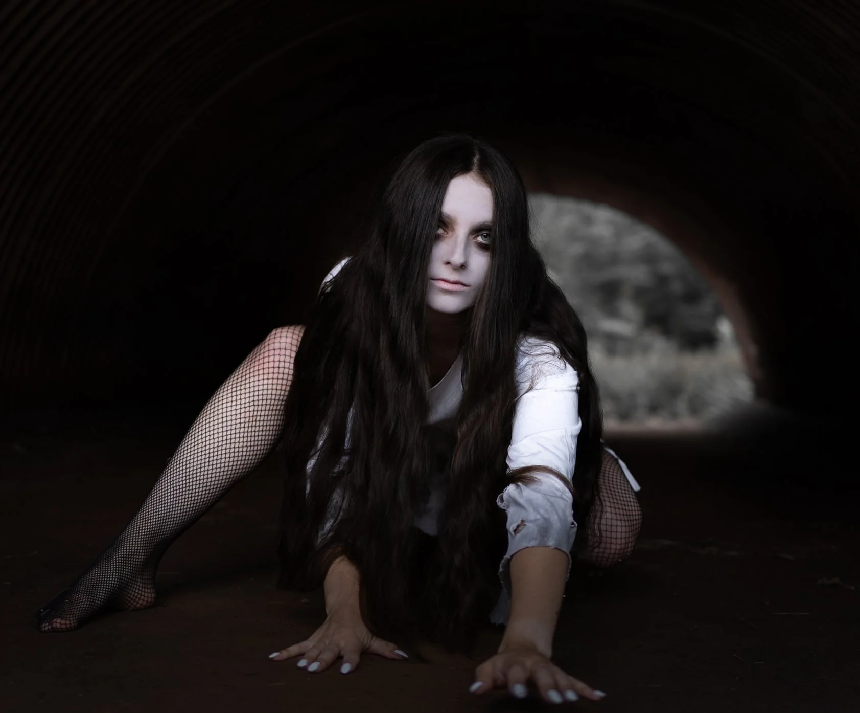 A woman with long dark hair, white face makeup, and patched white shirt crawling on the ground inside a dark tunnel.