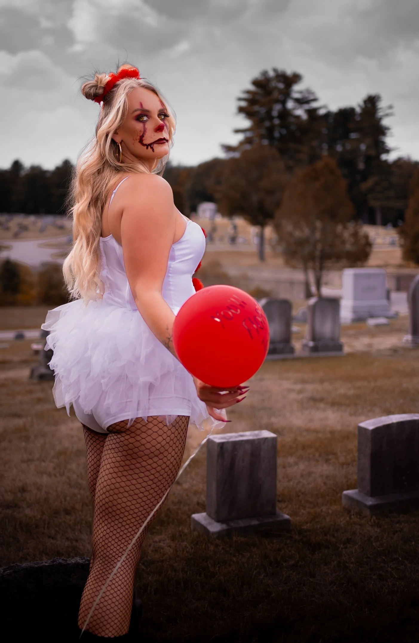 A woman dressed as a creepy clown with makeup and a tutu, holding a red balloon with writing, standing in a graveyard.