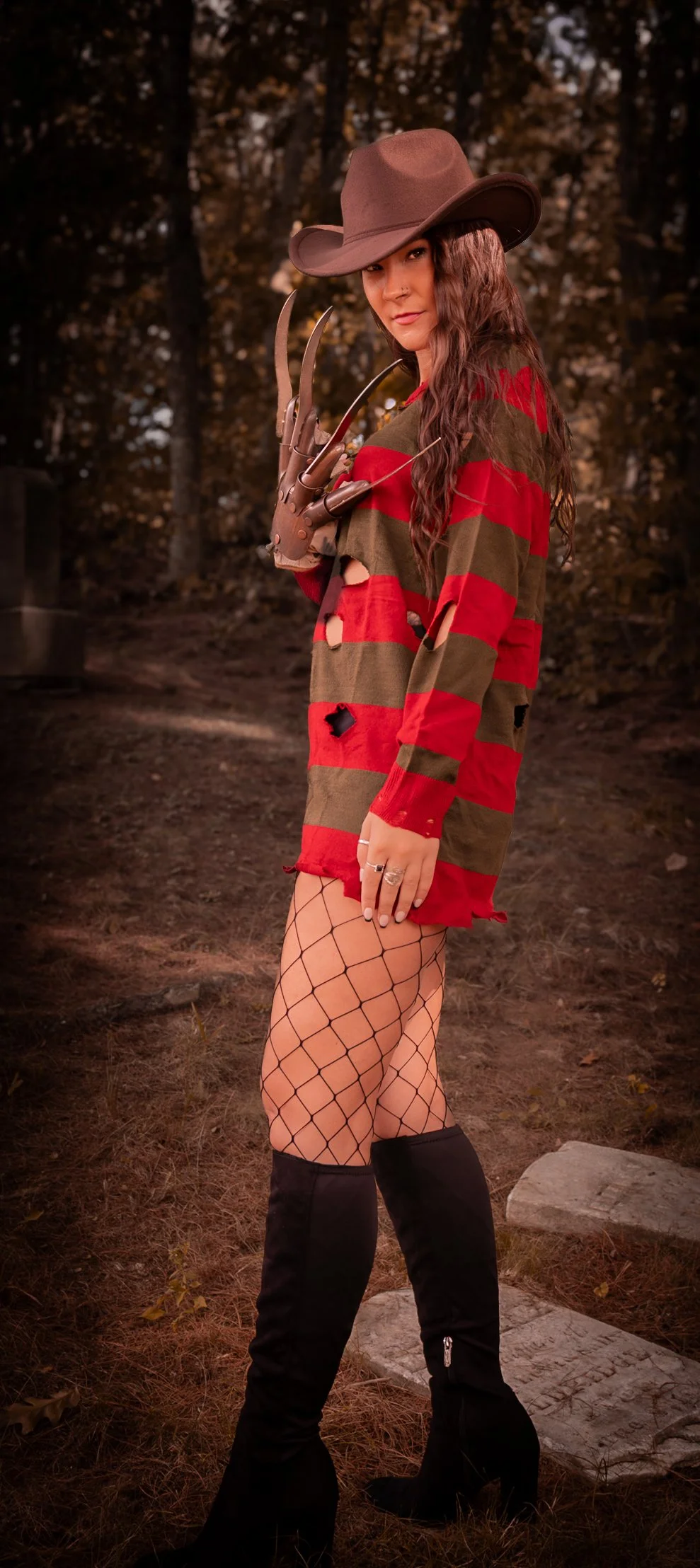 A woman dressed in a costume resembling Freddy Krueger from A Nightmare on Elm Street, standing outdoors in a wooded area with autumn trees. She wears a brown fedora hat, a red and green striped distressed sweater, fishnet stockings, and black thigh-
