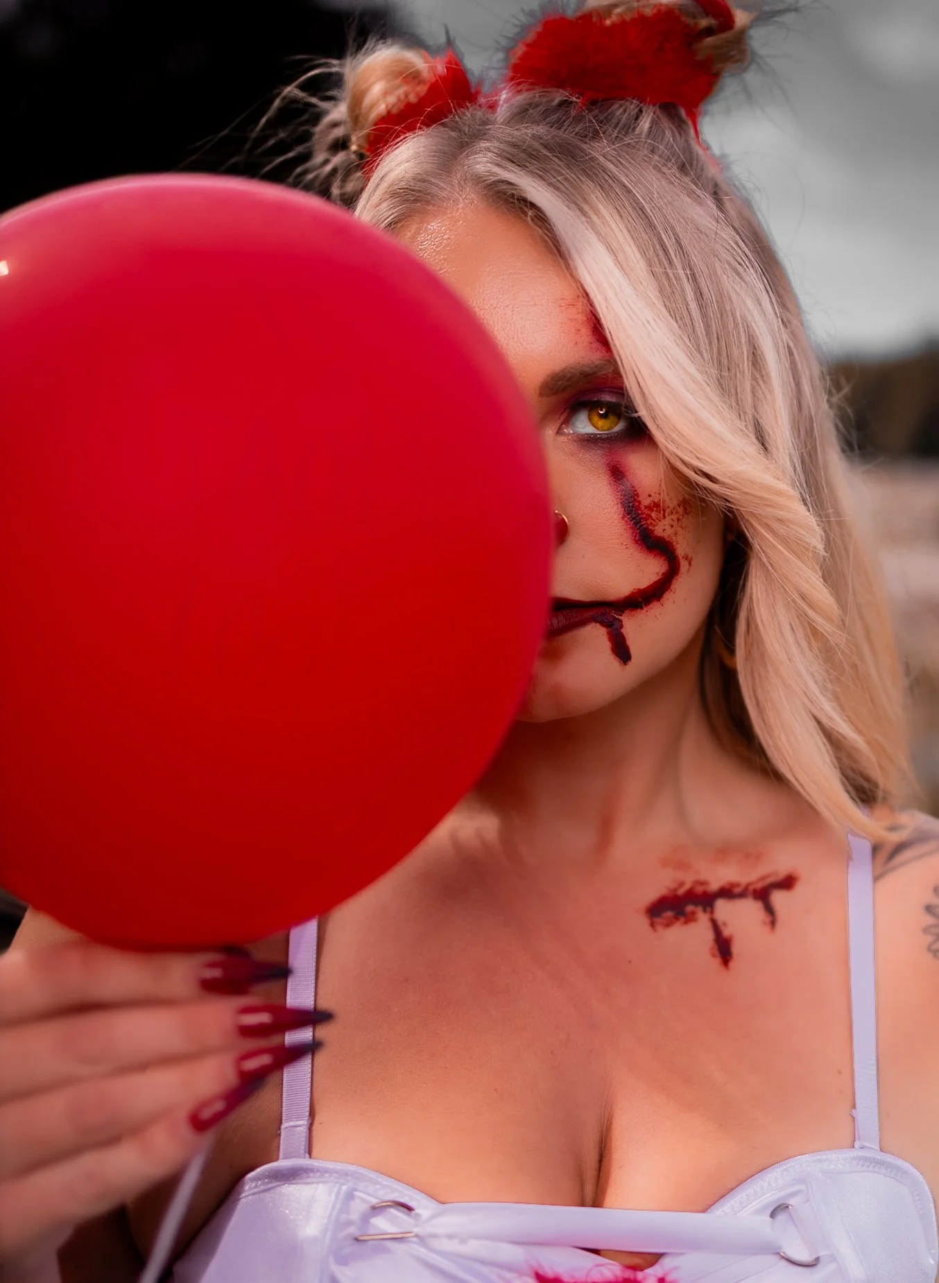 A woman with blonde hair, yellow eyes, and some face and shoulder makeup that looks like scars, holding a red heart-shaped balloon partially hiding her face.