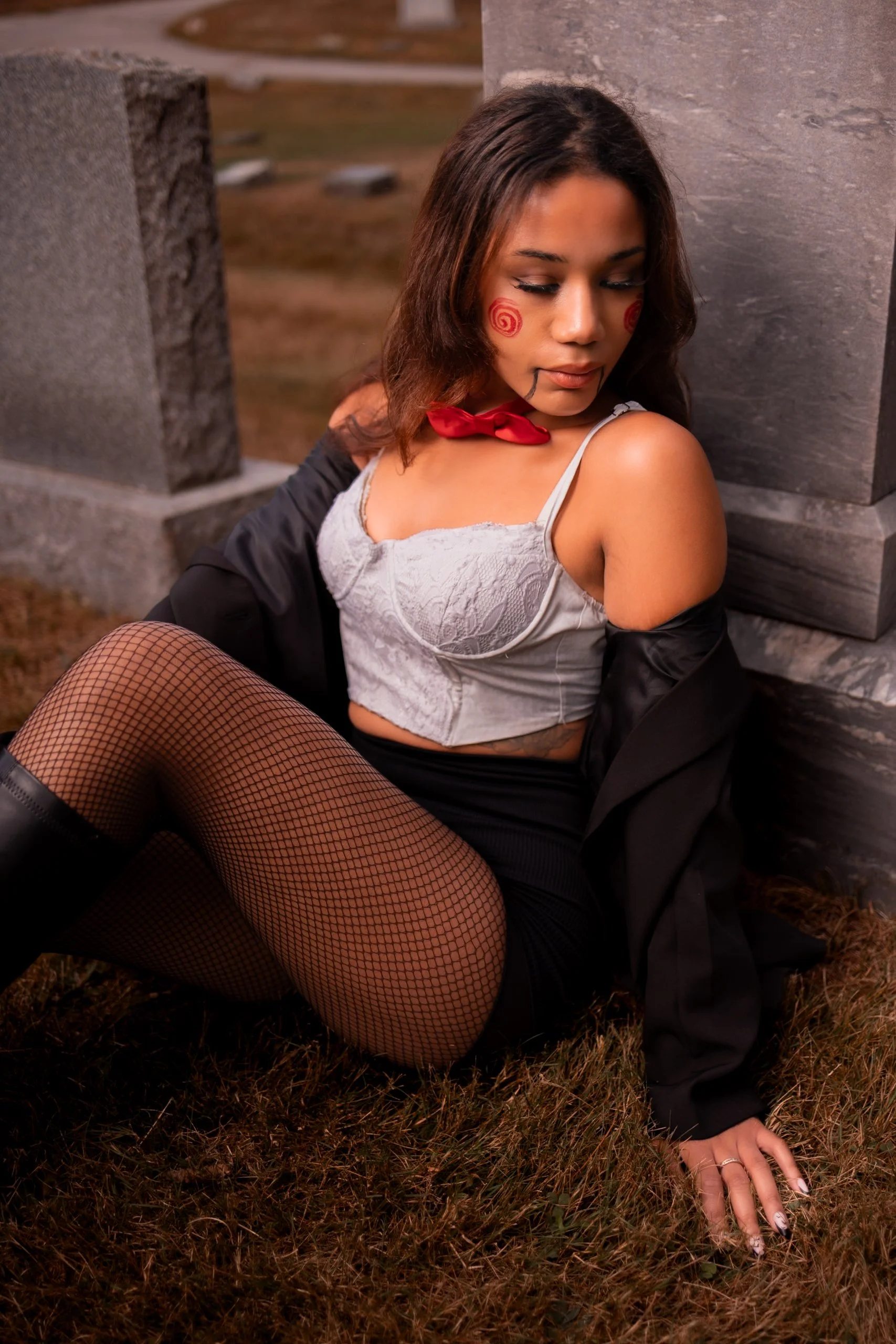 Woman with face paint, fishnet stockings, white lace corset, and black jacket sitting on grass against stone pillars.