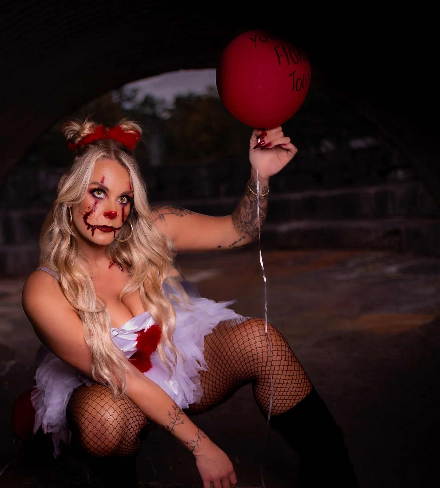 Woman dressed in a creepy clown costume with face paint resembling a doll, fishnet stockings, and a white tutu, holding a red balloon that says "You have a lovely smile today," under an arched stone bridge.