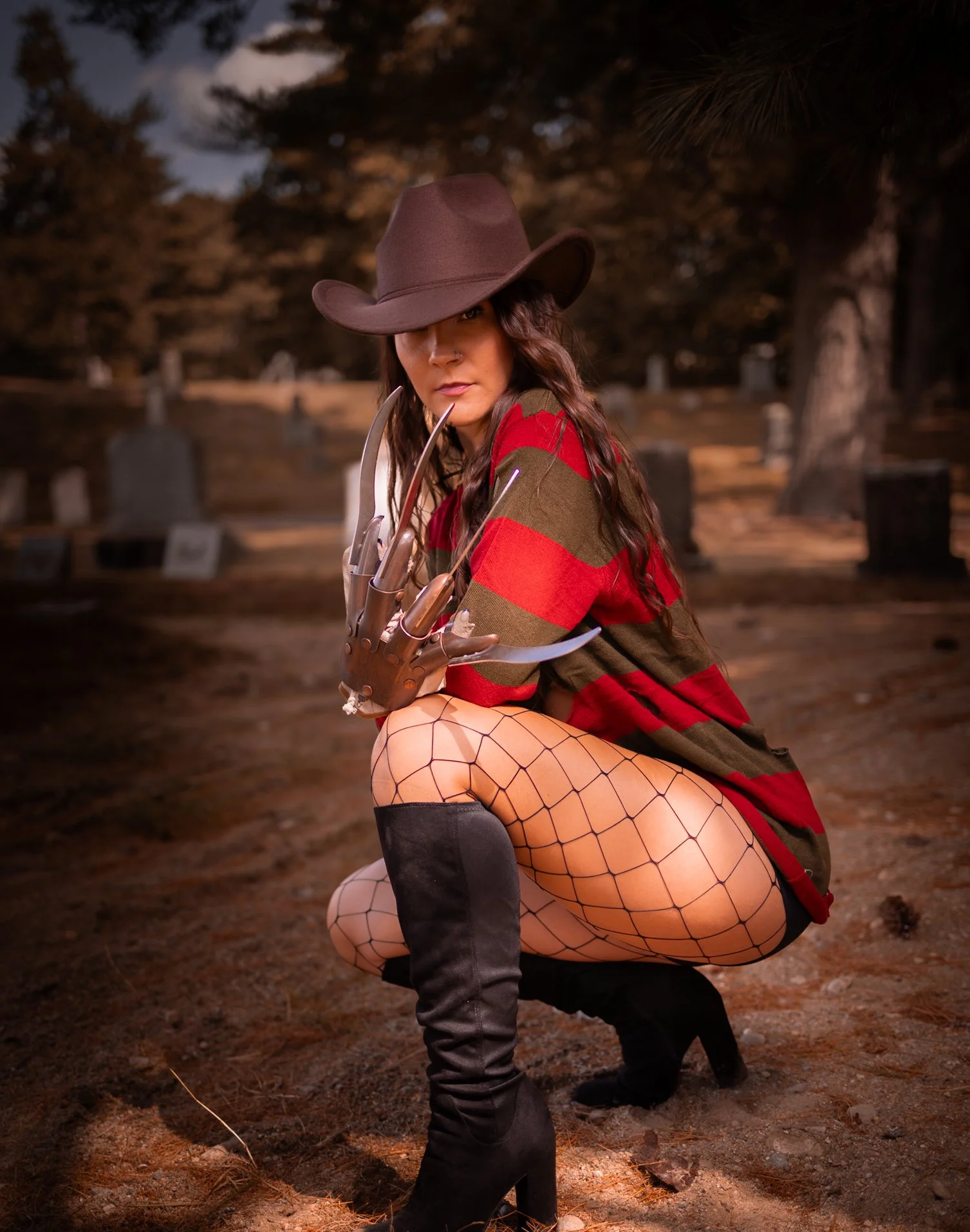 A woman dressed as a horror character resembling Freddy Krueger sitting on her knees in a graveyard at night. She wears a red and green striped sweater, fishnet stockings, black high-heeled boots, and a brown wide-brimmed hat. She has a metallic glov