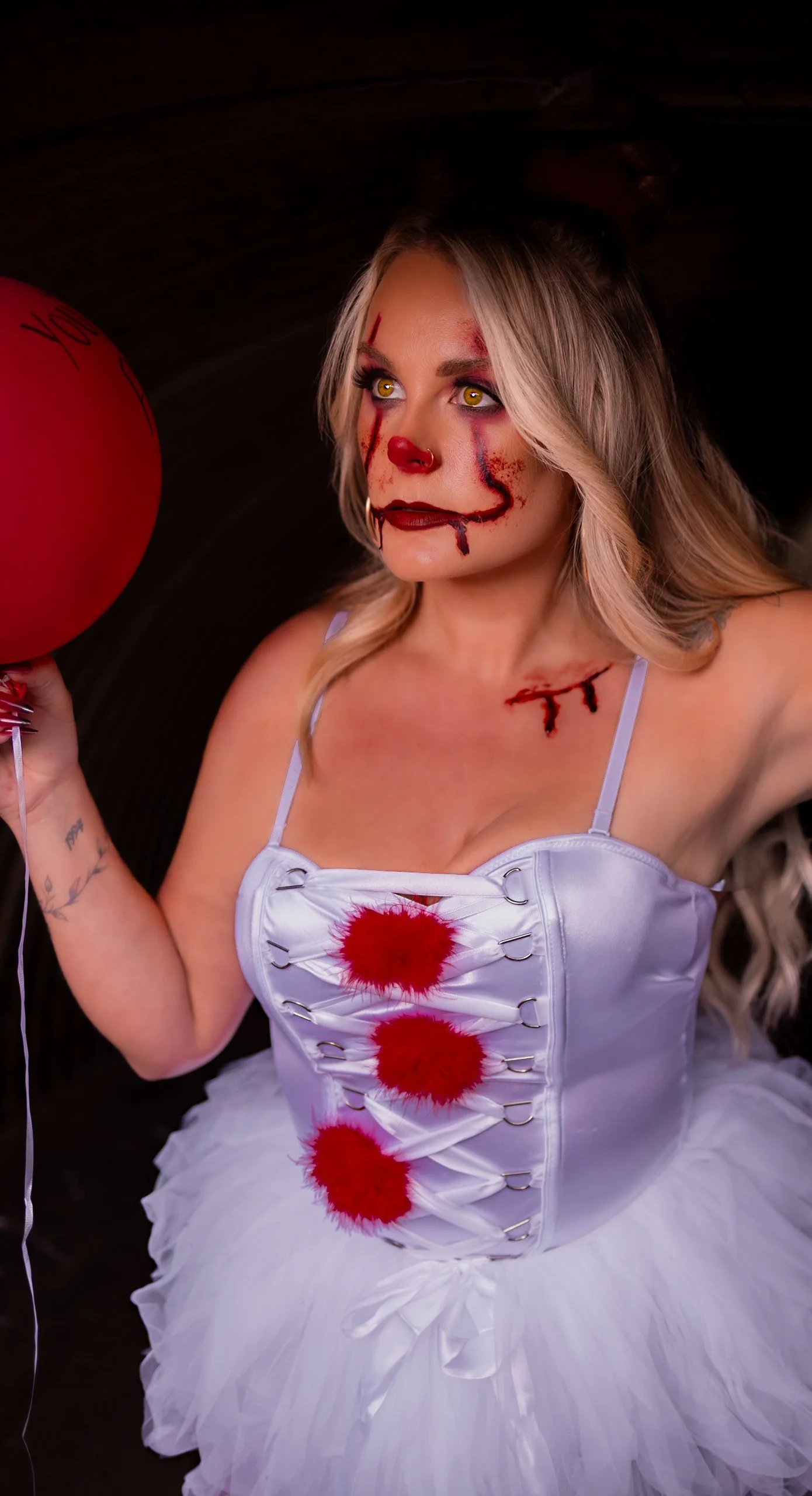 A woman dressed as a creepy clown or zombie with makeup resembling blood and wounds, holding a red balloon, wearing a white dress with red pom-poms and a tutu, in a dark setting.