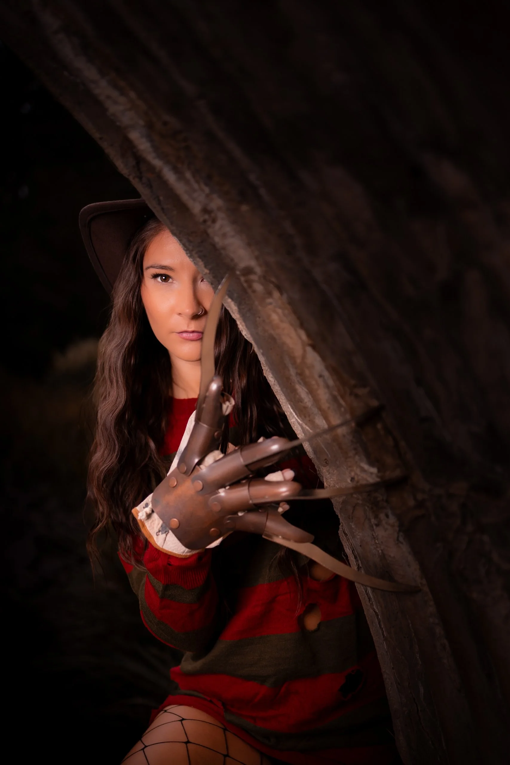 A woman with long wavy hair and a nose ring, wearing a red and black striped sweater and fishnet stockings, peeking through a narrow gap in a dark wooden surface, holding a metallic robotic hand.