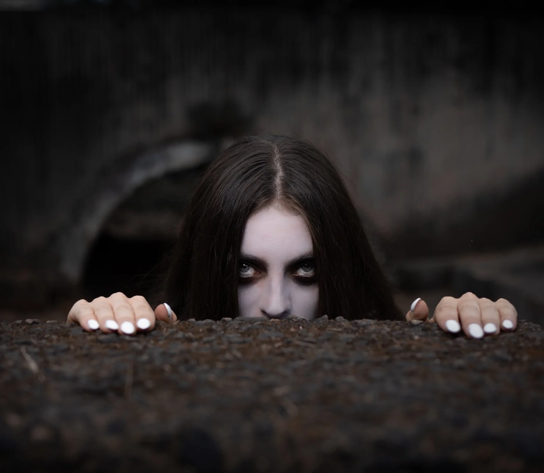 Woman with pale skin and dark hair peeking over dirt or rock, with intense eyes and hands gripping the surface