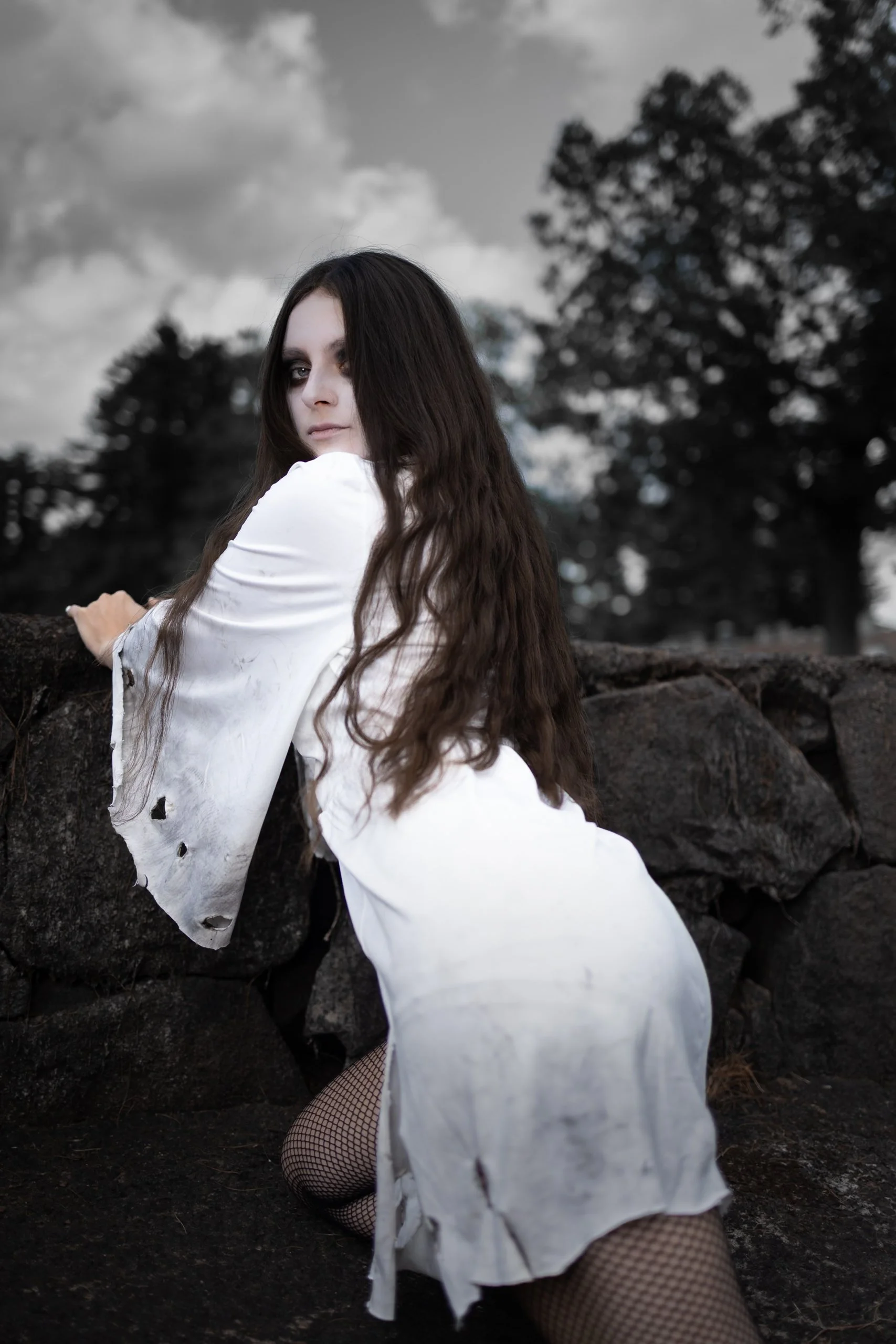 A woman with long dark wavy hair wearing a torn white dress and fishnet stockings, pose outdoors against a stone wall with trees and cloudy sky in the background.
