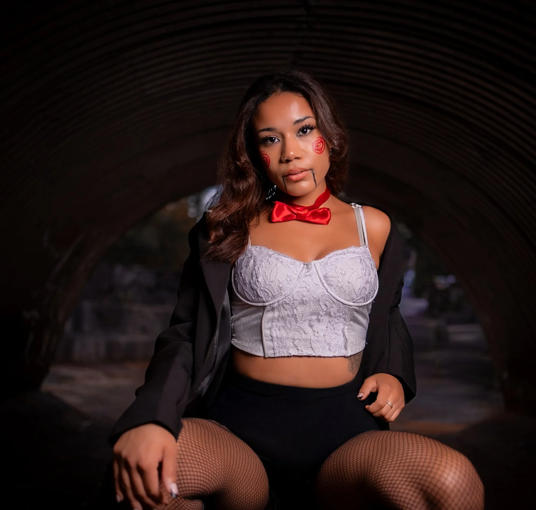 A woman with face makeup resembling a clown, wearing a lace crop top, fishnet stockings, a red bow tie, and a black jacket, sitting in a tunnel.