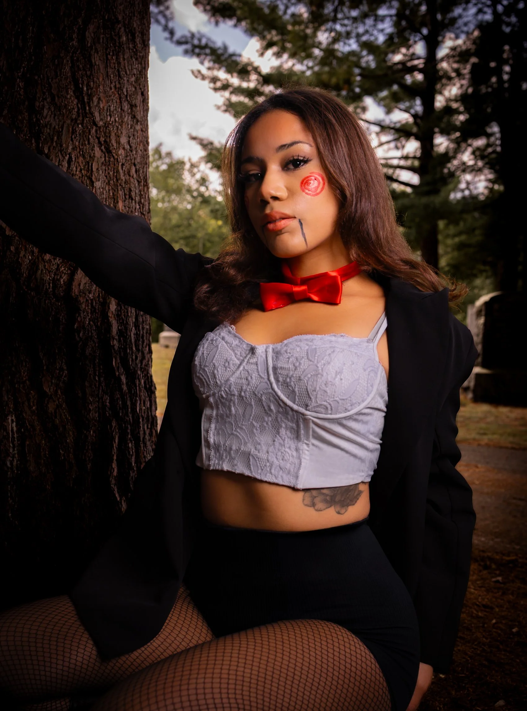 A woman with face paint resembling a clown, wearing a red bow tie, a white lace crop top, black jacket, fishnet stockings, and black shorts, leaning against a tree in a forested area.