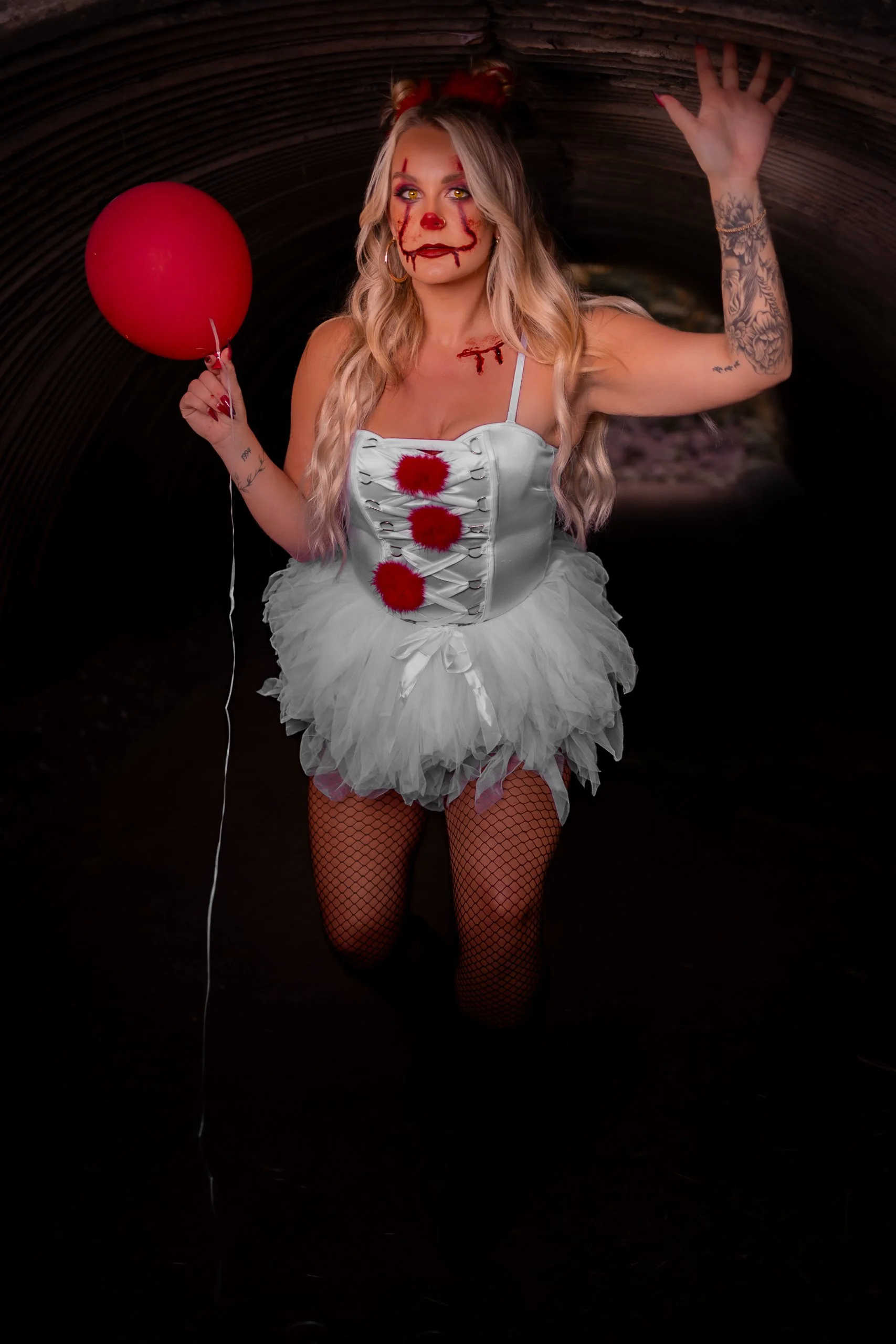 Person dressed in a clown costume with face paint resembling a clown, holding a red balloon, inside a dark tunnel.