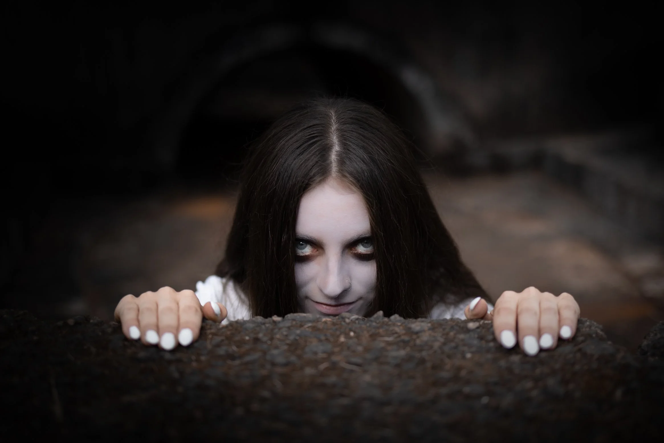 A woman with dark hair and pale face crawling out of a dark tunnel, looking directly at the camera with intense eyes.