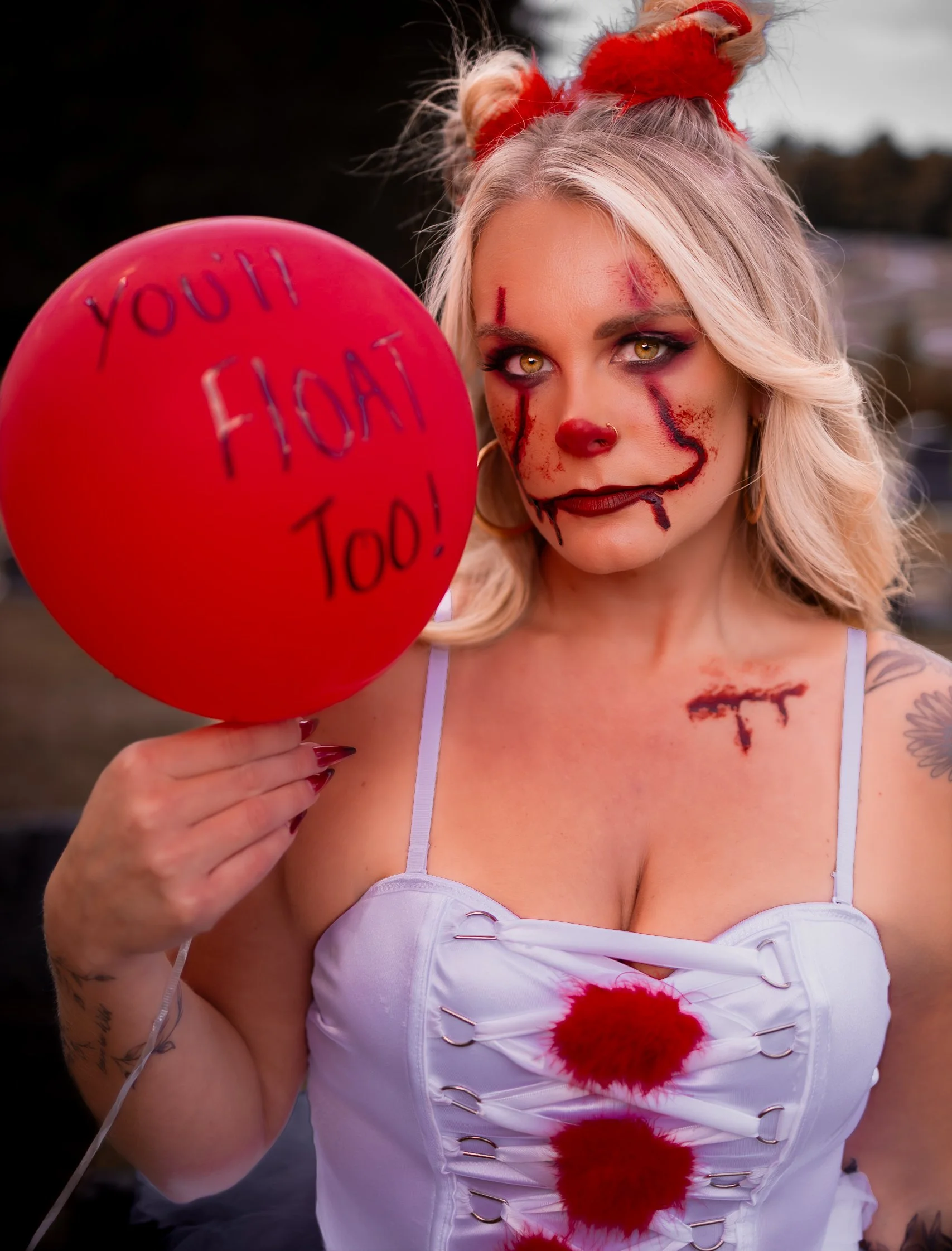 Woman dressed as a creepy clown holding a red balloon with 'You' and 'Too' written on it. The woman has blonde hair, two buns with red ribbons, and makeup with a fake bloody smile, scars, and dark eyes.