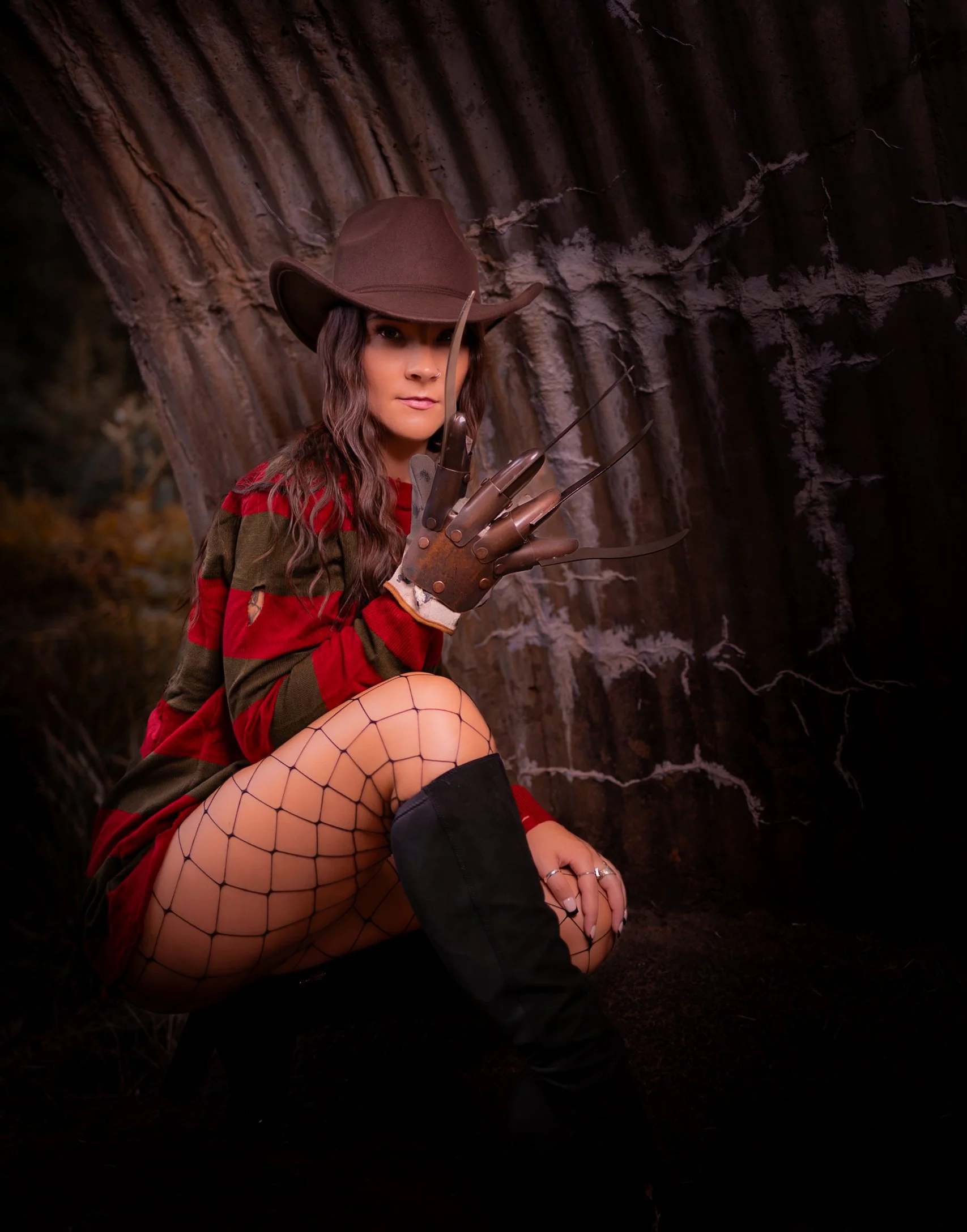 Woman dressed as horror character with a hat, striped sweater, fishnet stockings, and leather glove with claws, crouching in front of a large tree trunk.