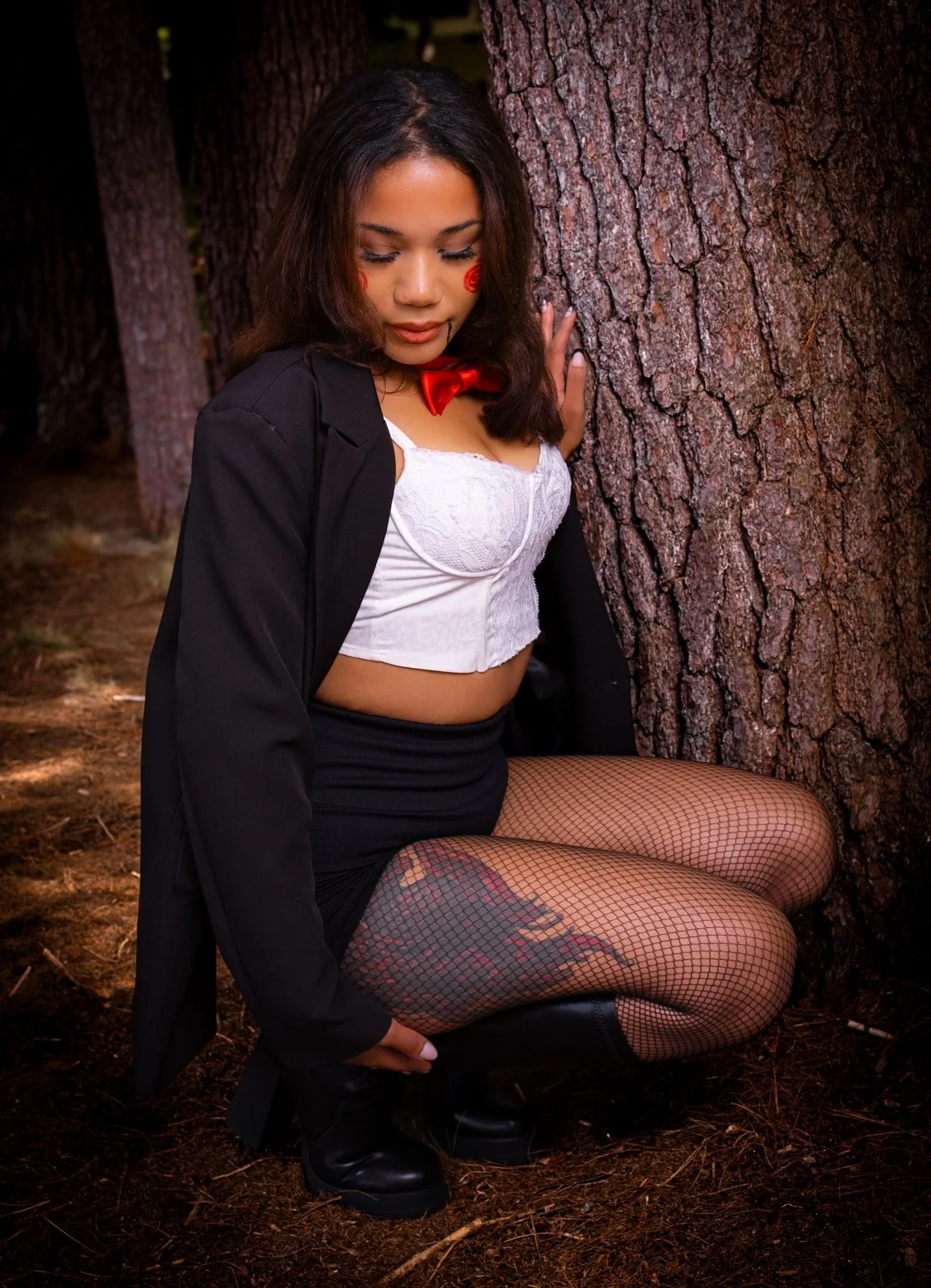 A woman kneeling by a tree in a forest, wearing fishnet stockings, a white crop top, black skirt, black boots, a black blazer, and a red bow tie, with Halloween face paint and curly brown hair.