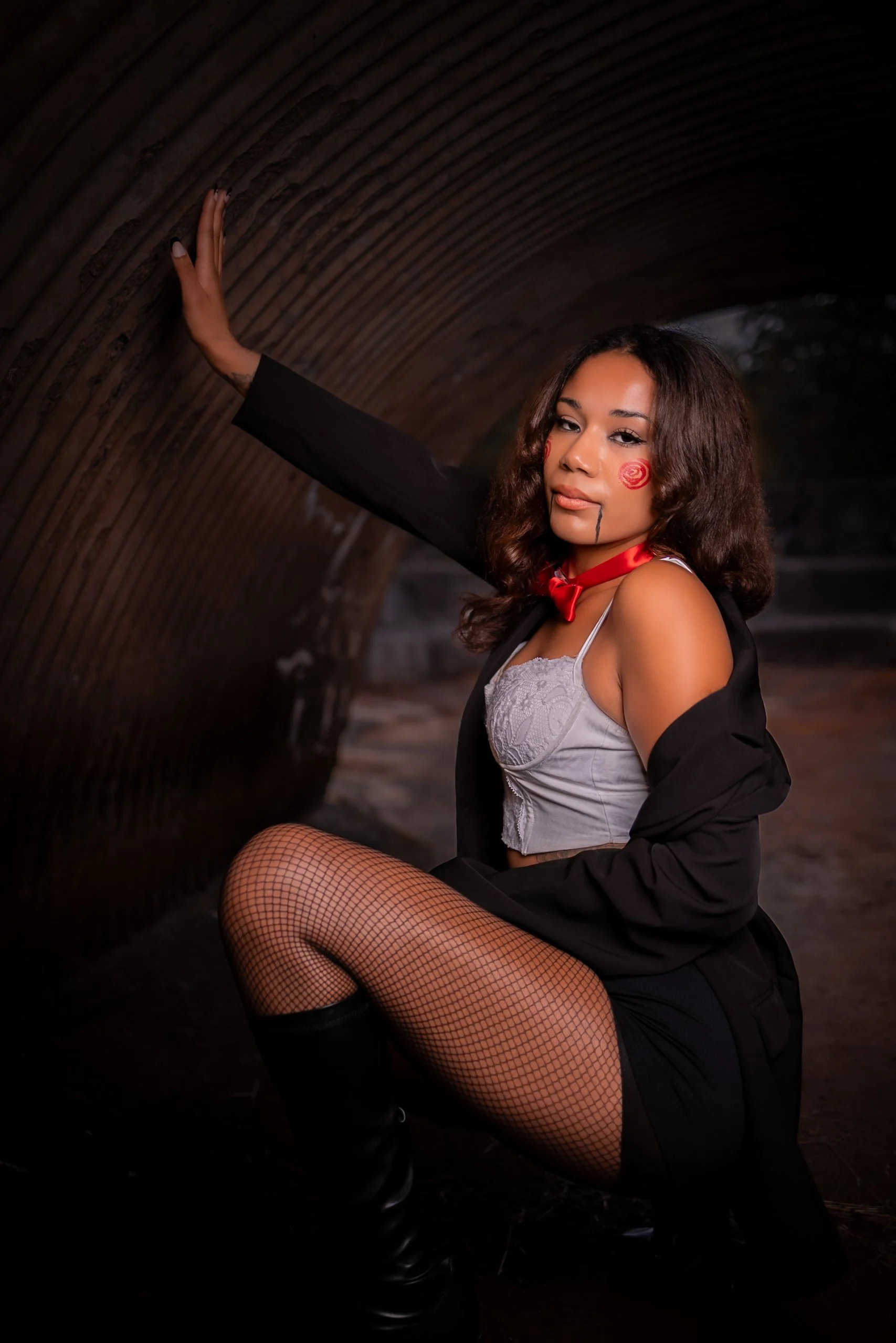 A woman dressed in a schoolgirl costume with makeup, fishnet stockings, and a black jacket posing in a tunnel.
