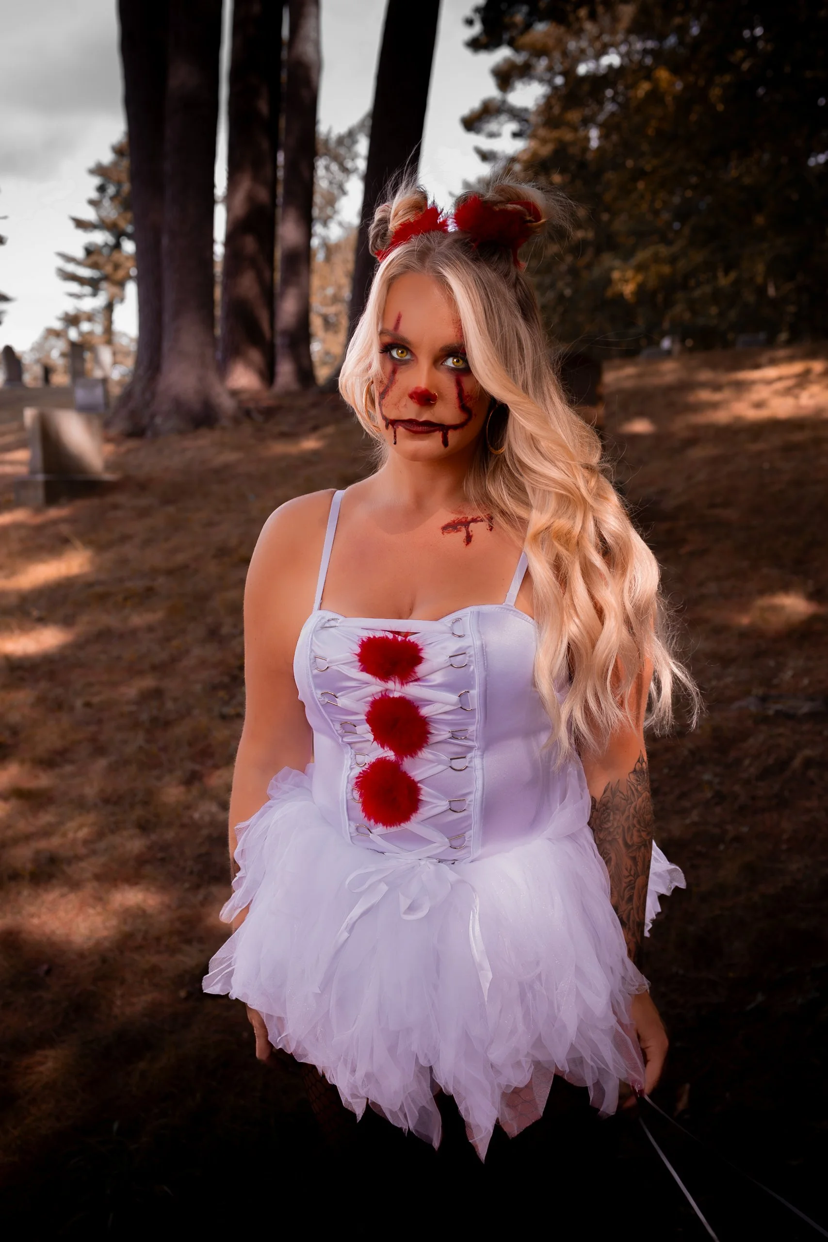 A woman dressed in a Halloween costume with face makeup resembling a creepy clown or doll, featuring blood-like streaks and a stitched mouth. She has long, blonde wavy hair adorned with red furry hair accessories. She is wearing a white dress with re