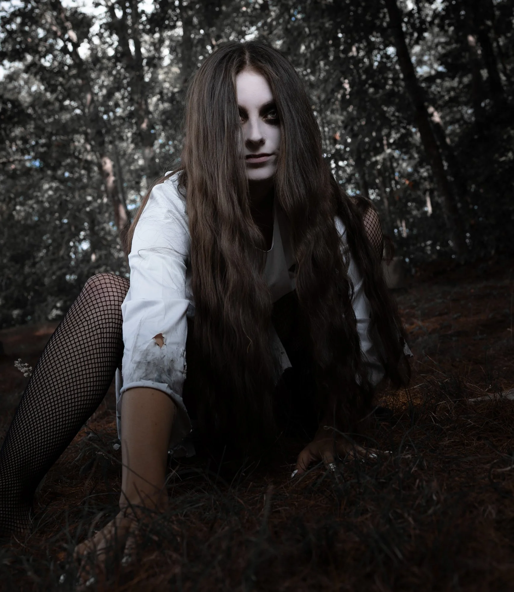 A young woman with long dark hair and dark makeup kneels on the ground in a wooded area, wearing a torn white shirt and fishnet stockings, with a dark, eerie expression.