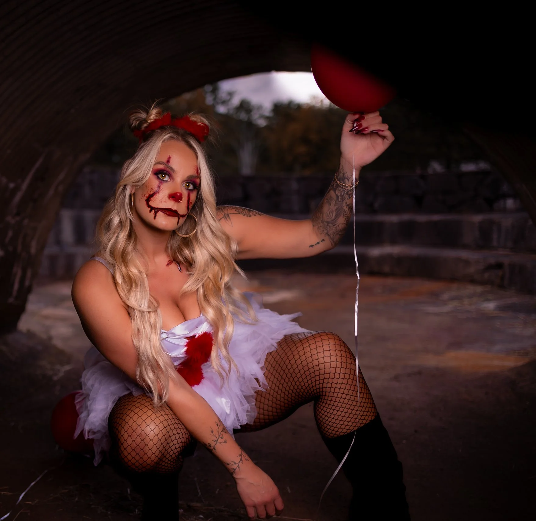 A woman dressed as a creepy clown crouches under a bridge holding a red balloon, with makeup that looks like a clown with scars, in a Halloween costume.