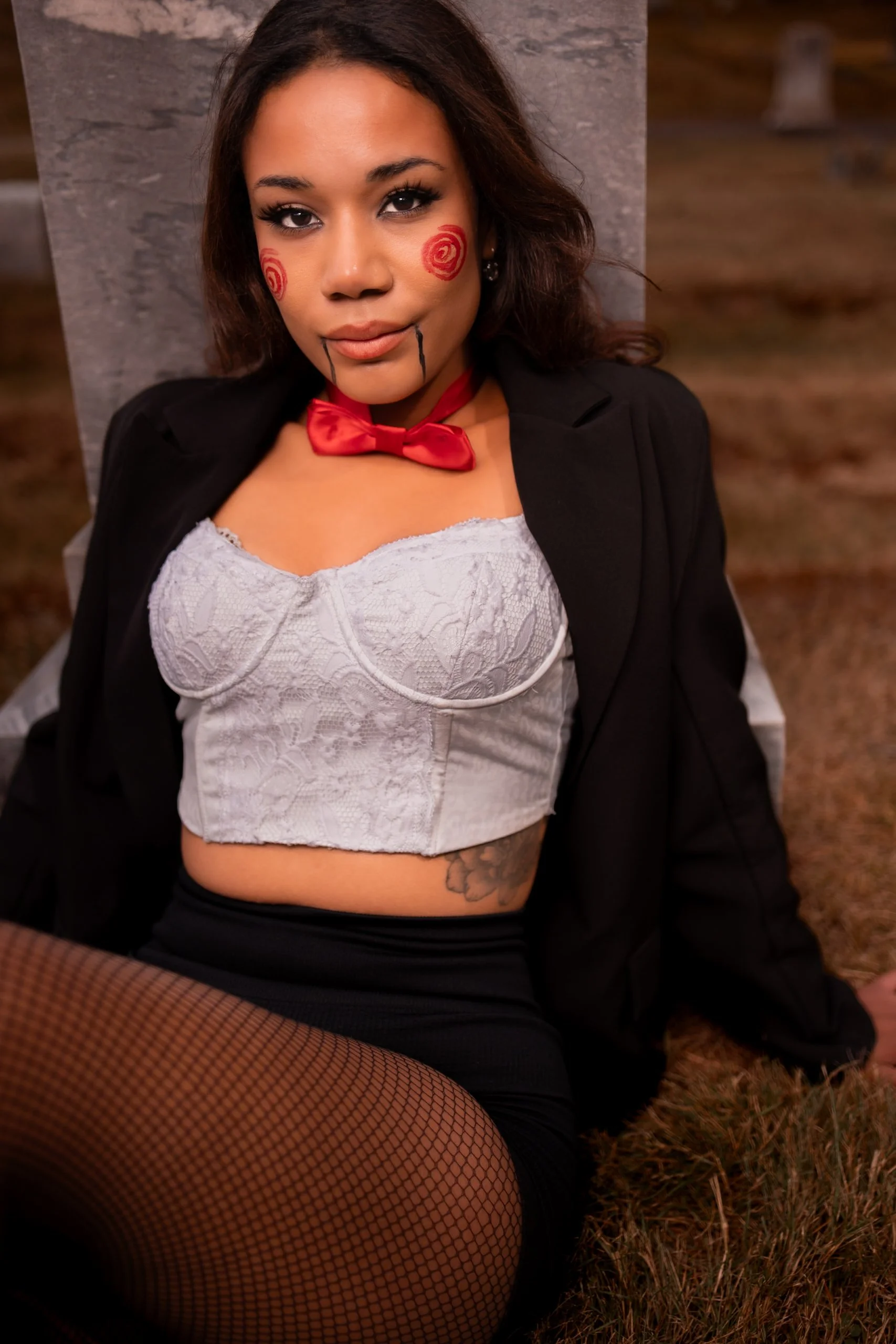 A woman sitting outdoors on the grass, wearing a black blazer, white lace crop top, fishnet stockings, and a red bow tie. She has face paint resembling red swirls on her cheeks and chin, and her makeup and hair are styled elaborately.