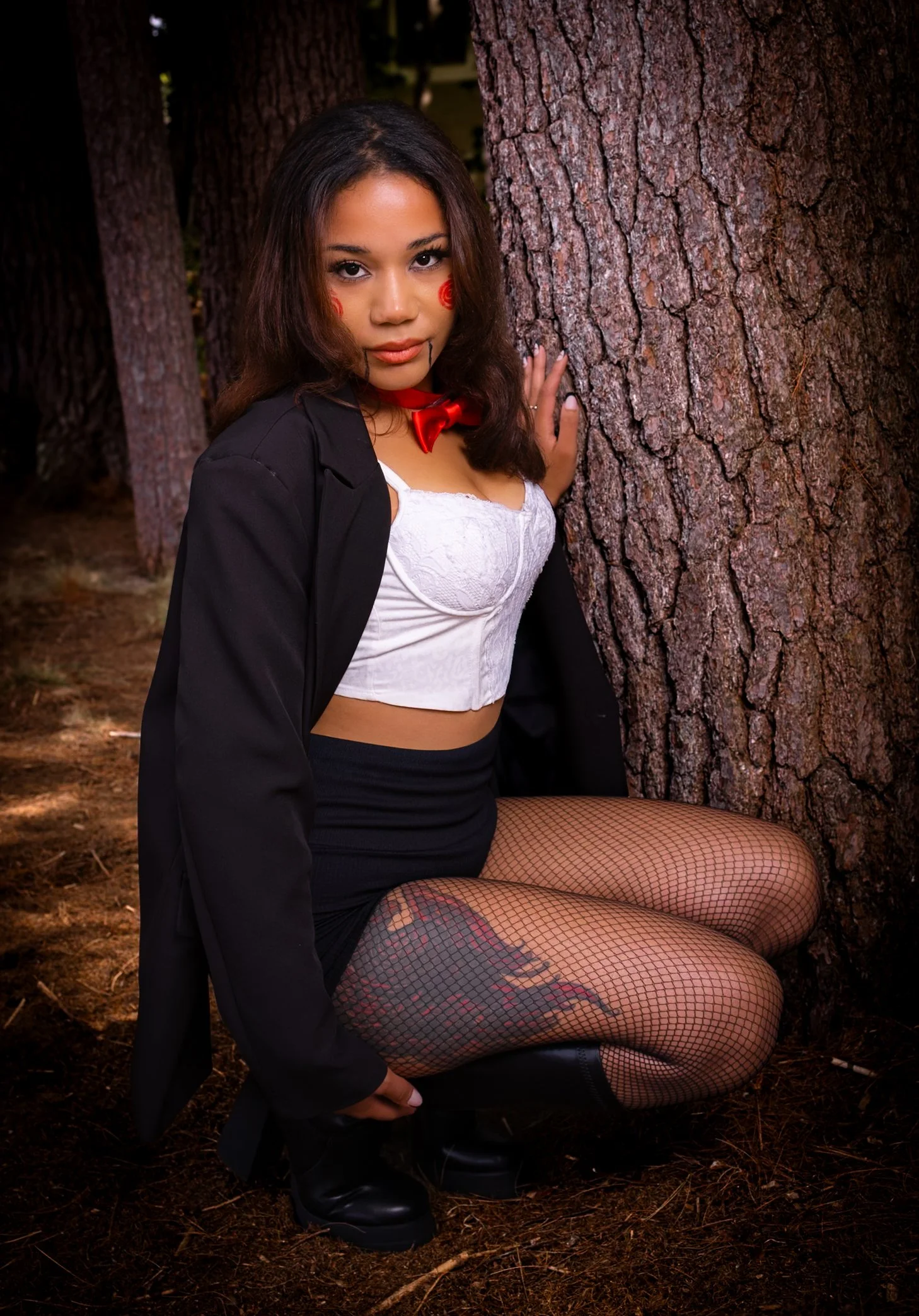 A young woman with dark hair and makeup sitting on the ground in a wooded area, touching a tree trunk, wearing a white lace crop top, black skirt, black jacket, fishnet stockings, combat boots, red lipstick, and face paint, with a red bow tie around 