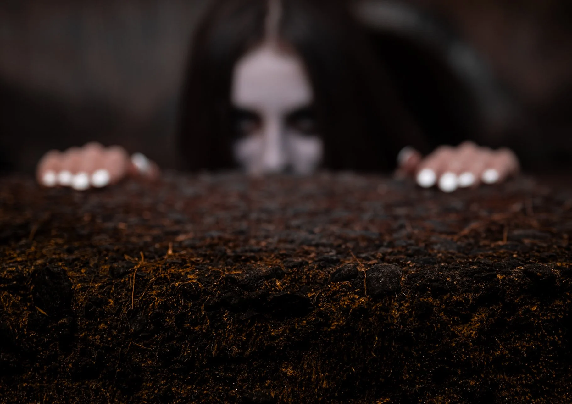 A woman with dark, loose hair and dark makeup peering over a rough, textured surface, with only her eyes and hands visible.