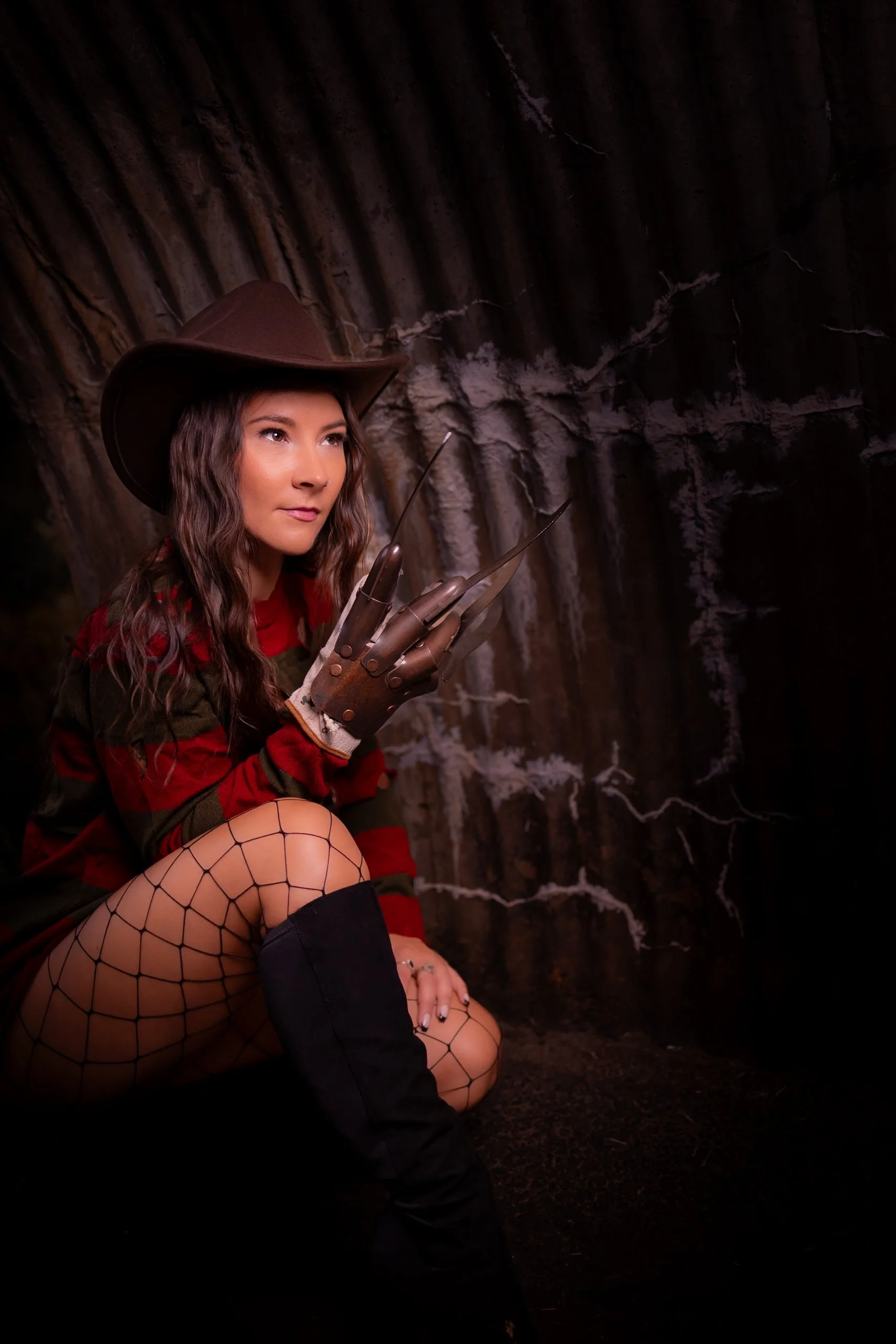 A woman dressed as Freddy Krueger with a cowboy hat and fishnet stockings crouched inside a dark, rocky cave, wearing a glove with metal blades.