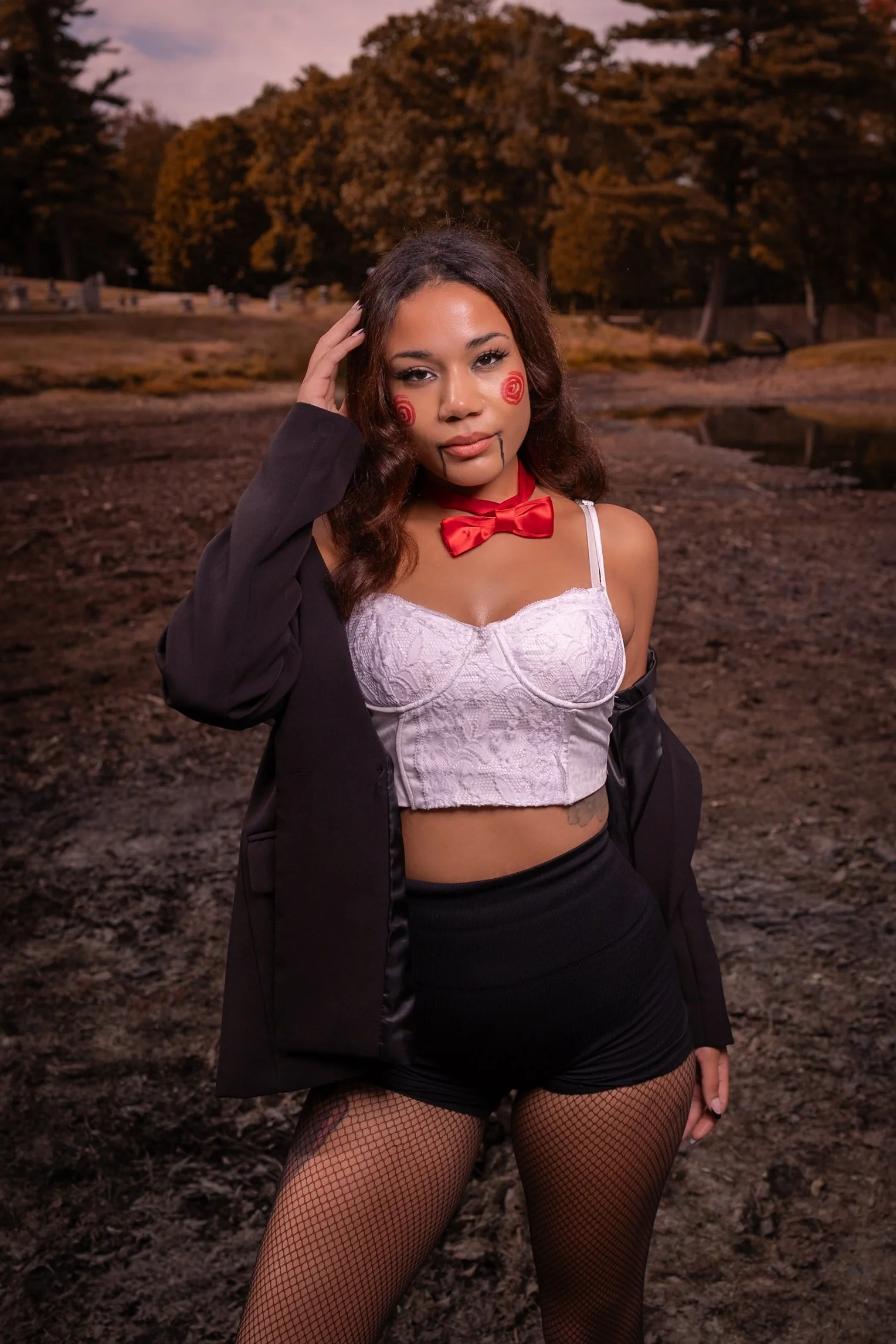 A woman dressed as a horror movie character with gory makeup, black jacket, white lace crop top, fishnet stockings, and a red bow tie, standing outdoors near a pond with trees in the background.