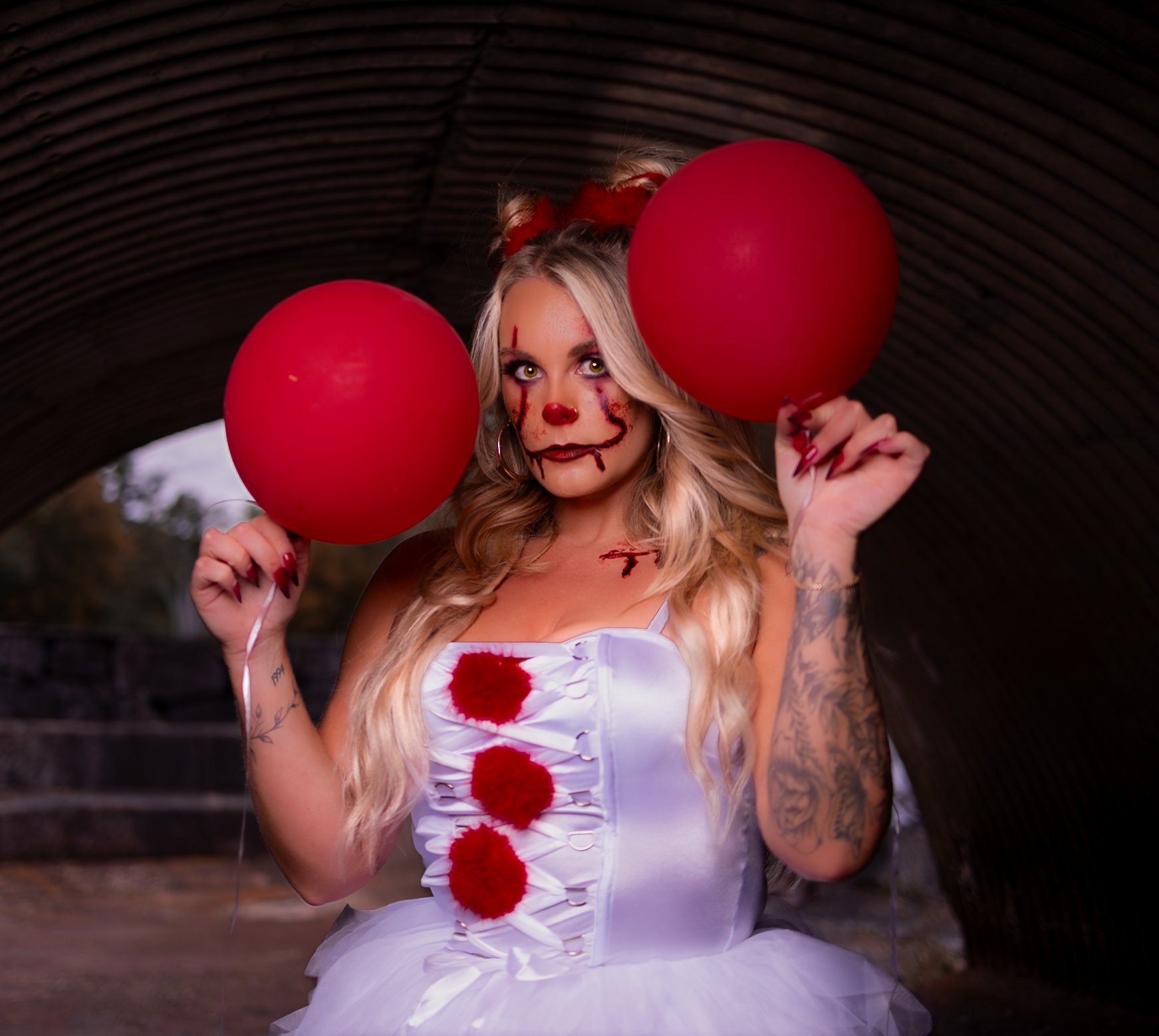 Woman with clown makeup, blonde hair, wearing a white dress with red pom-poms and holding two red balloons, standing under a bridge.