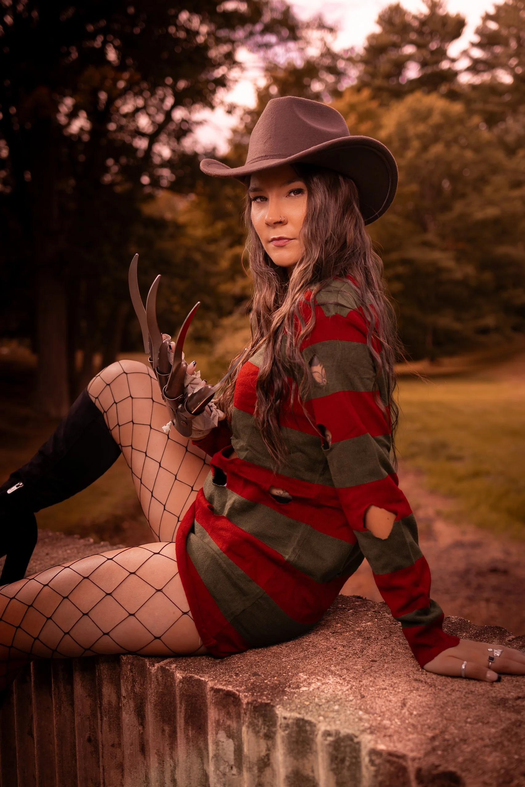 A woman dressed as Freddy Krueger from Nightmare on Elm Street, wearing a brown fedora, red and green striped sweater with holes, fishnet stockings, and holding a clawed glove, sitting outdoors on a rock with a forested background at sunset.