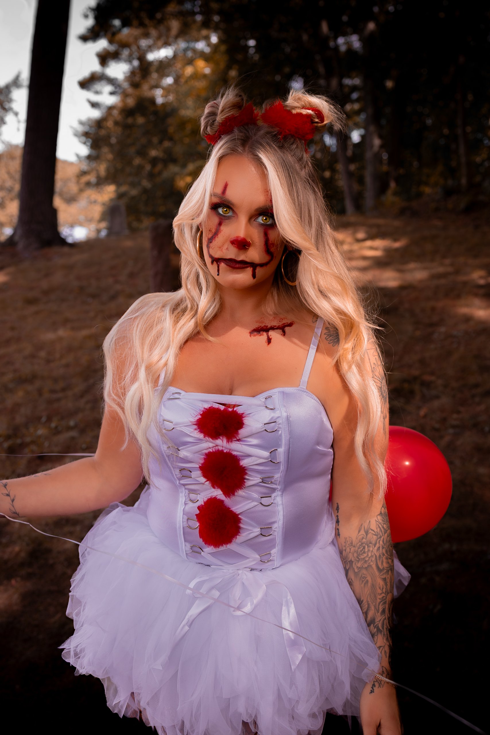 Woman dressed in a white dress with red fake blood and horror makeup, outdoors with trees and a red balloon in the background.