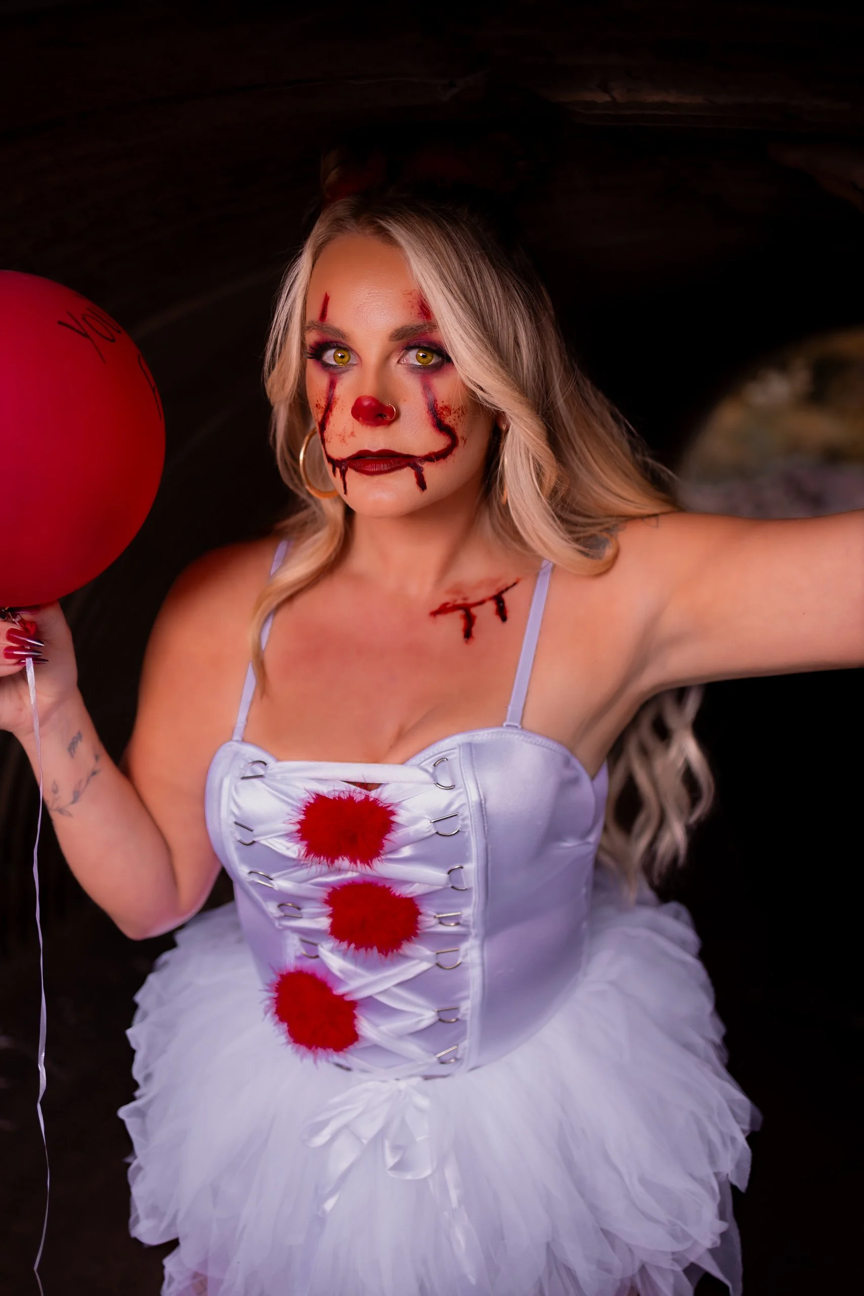 Woman with clown makeup and bloodstains, holding a red balloon, inside a dark tunnel.