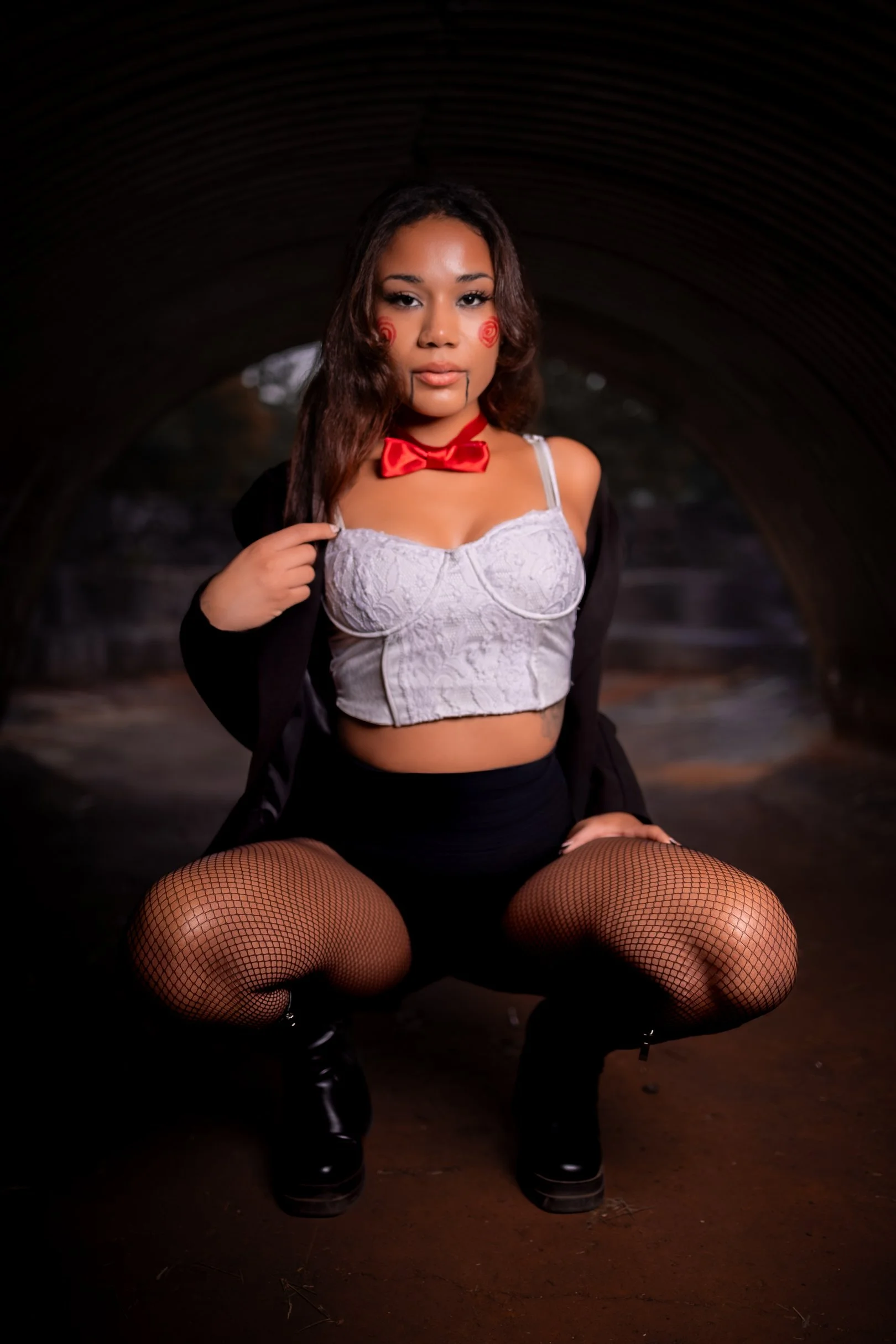Young woman crouching under a tunnel, wearing lingerie with fishnet stockings, black boots, red bow tie, and clown makeup with red circles on cheeks, dark eye makeup, and lipstick.