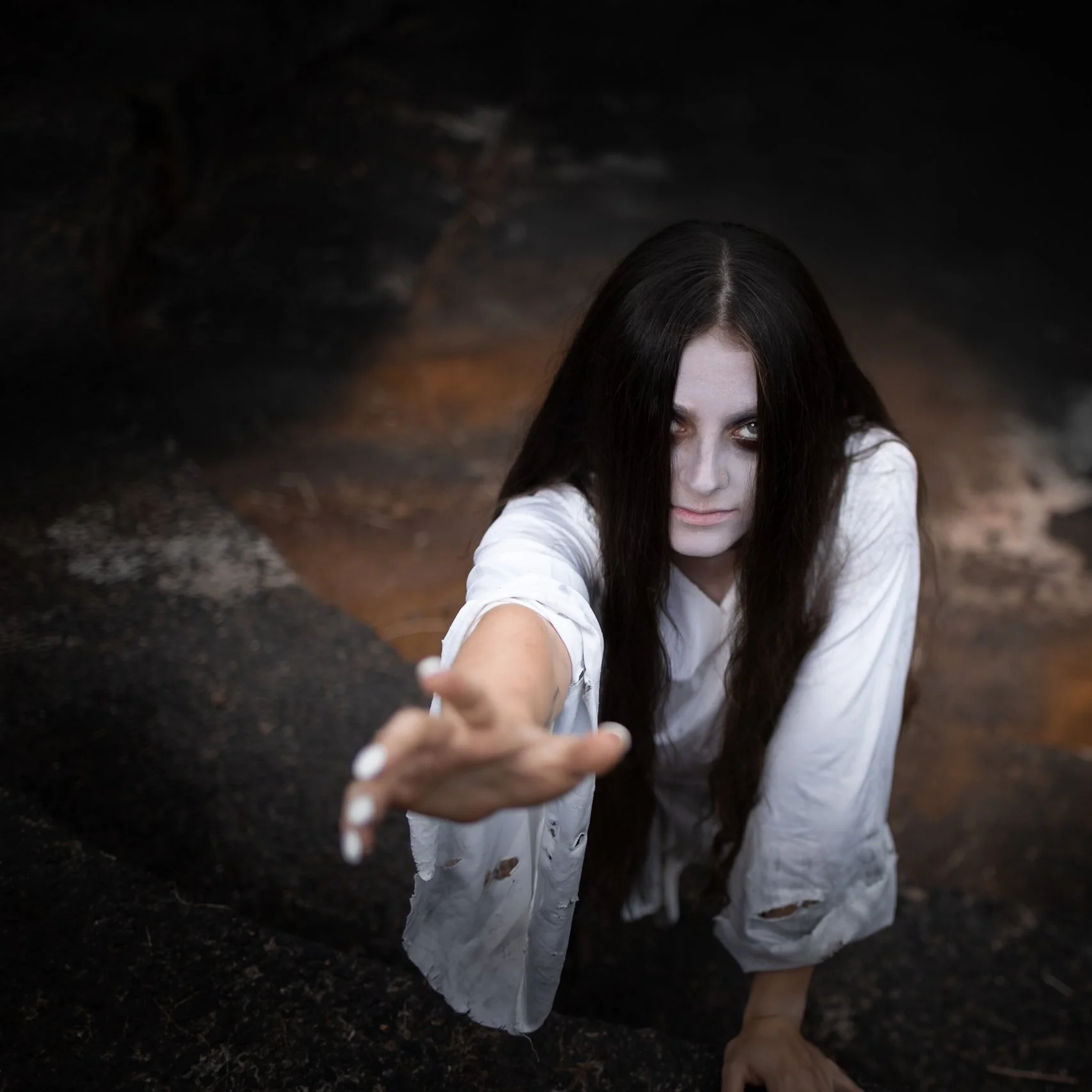 A woman with long dark hair and pale makeup reaching out with her hand, wearing a white, slightly torn shirt, in a dark, cave-like environment.