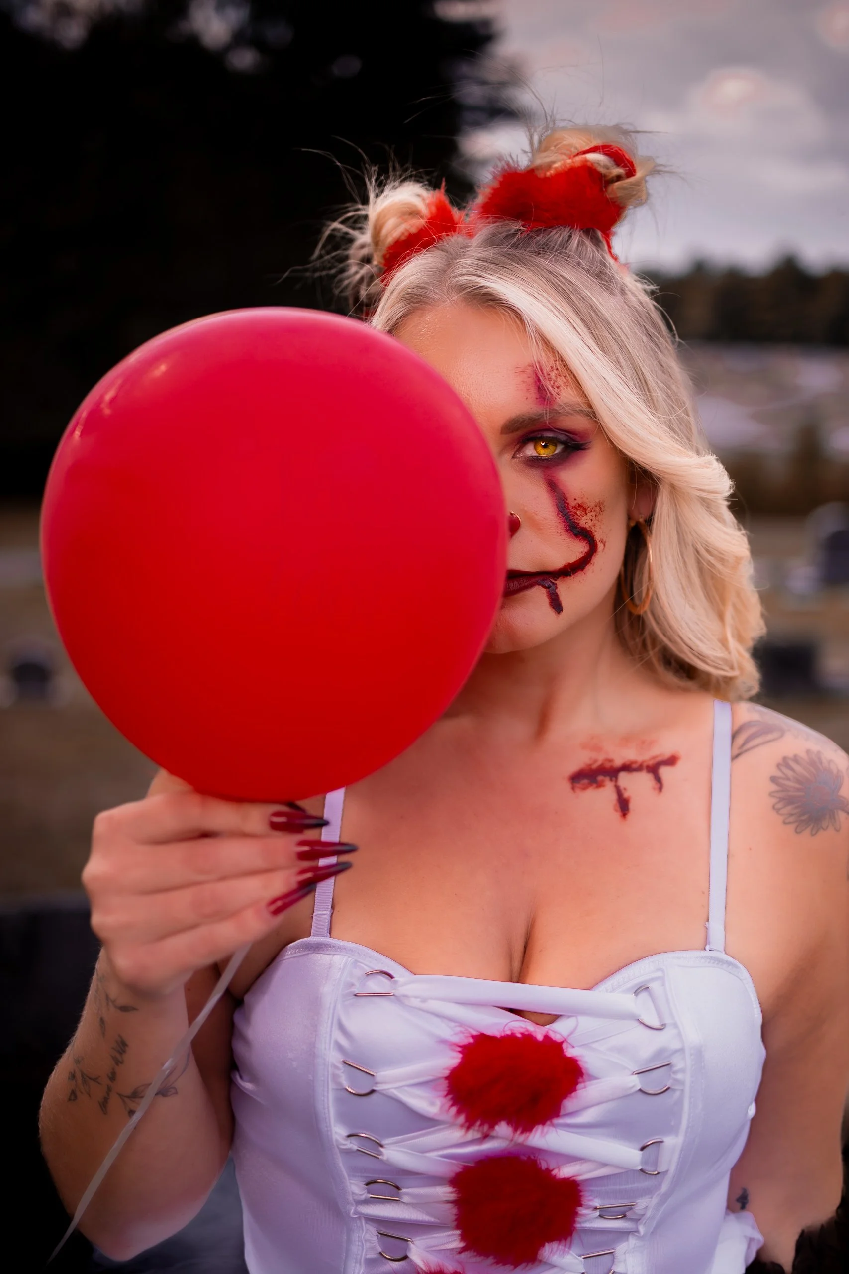 A woman with Halloween makeup and scars holds a red balloon in front of her face outdoors, with a dark sky and trees in the background.