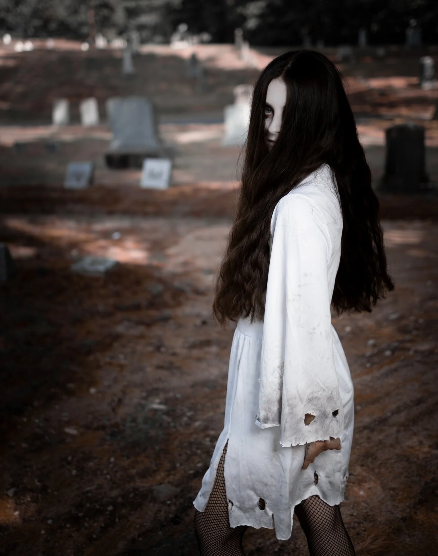 A woman with long dark hair and pale face wearing a torn white dress and fishnet stockings stands in a graveyard at dusk, looking over her shoulder with a sinister expression.