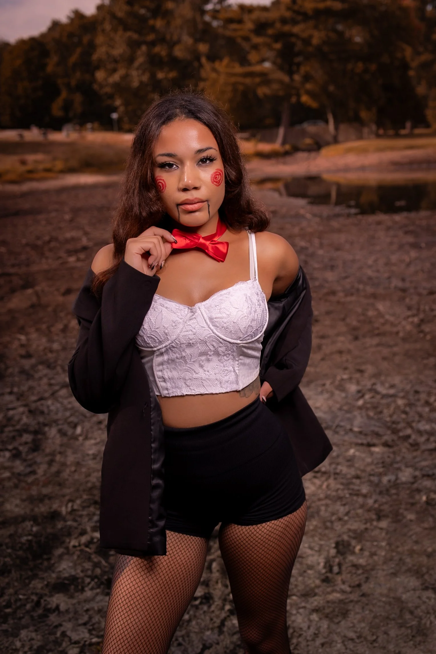 A young woman dressed in a horror clown costume with face paint, a white lace crop top, a black jacket, black shorts, fishnet stockings, and a red bow tie, standing outdoors on a dirt ground with trees and a small pond in the background during sunset