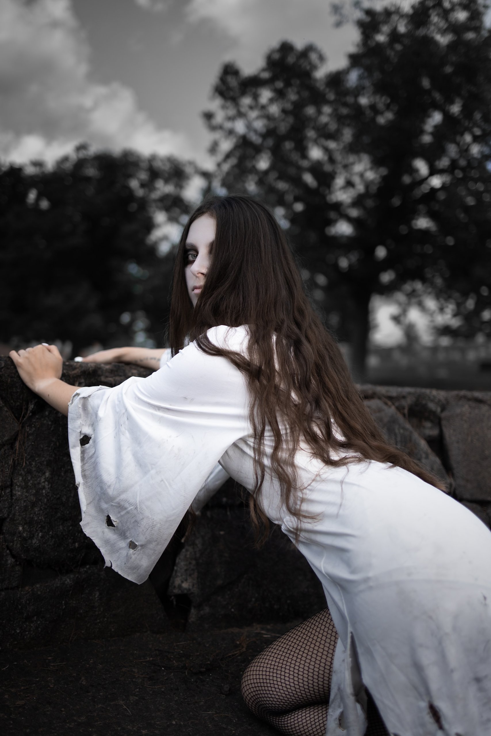 A woman with long dark hair, wearing a torn white dress, kneeling on the ground with her hands on rocks, in an outdoor setting with trees and a cloudy sky in the background.