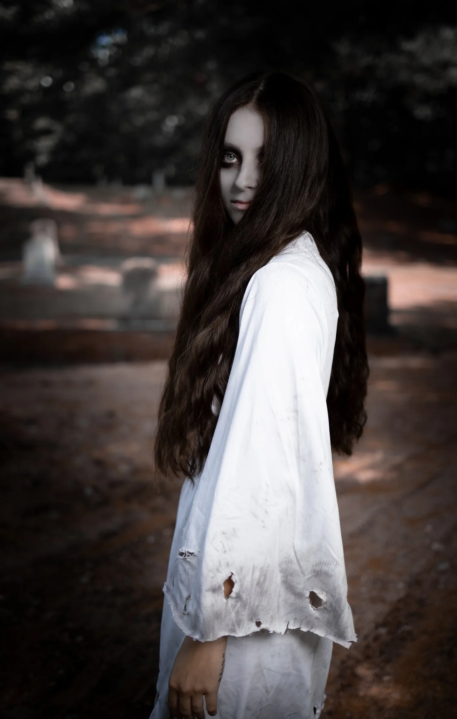 A young woman with long dark hair and pale complexion in a torn white shirt standing in a dark, spooky outdoor setting.