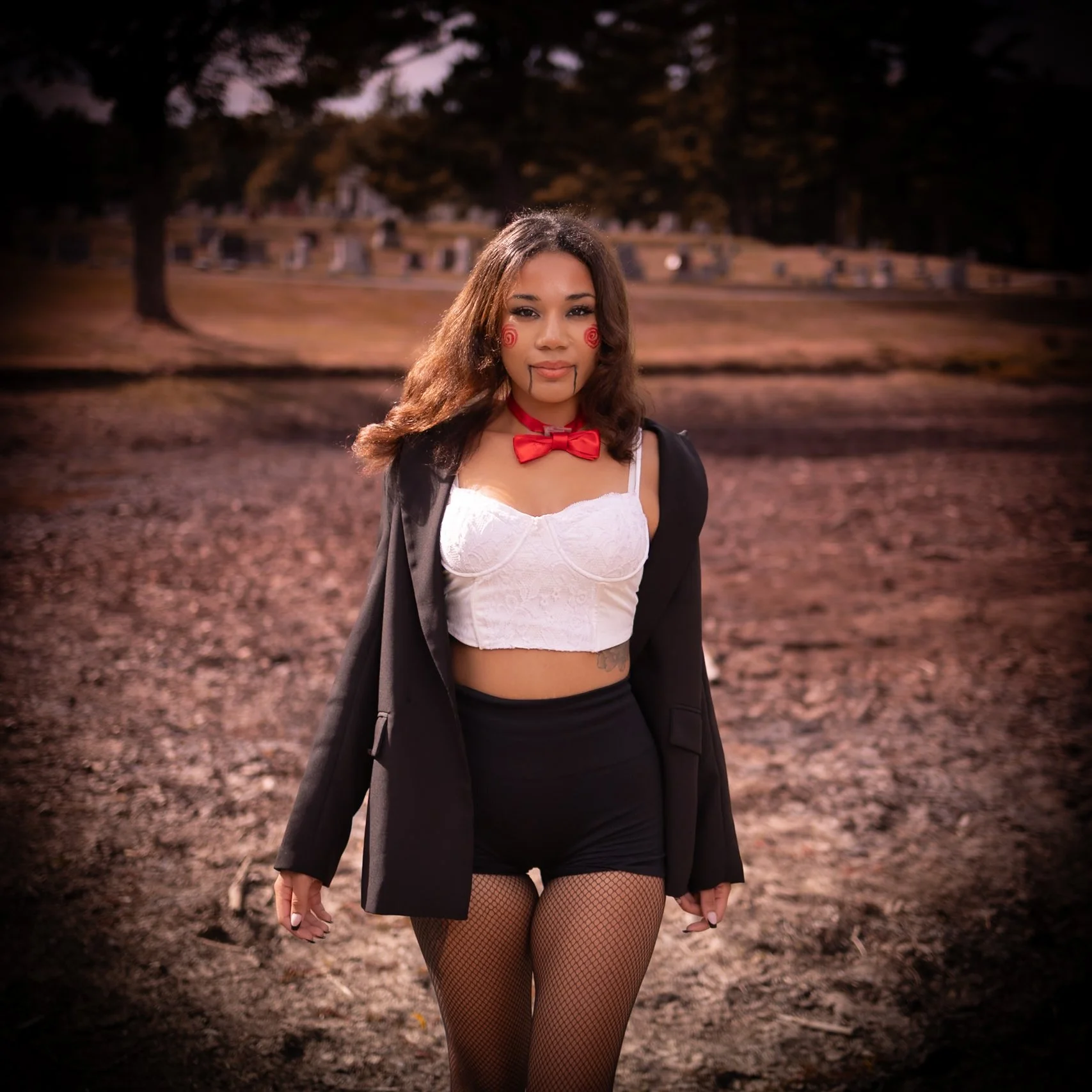Young woman standing outdoors in costume, with face paint and makeup resembling a doll or clown, wearing a white cropped top, black shorts, fishnet stockings, a red bow tie, and a black blazer draped over her shoulders, on a dirt path with trees and 