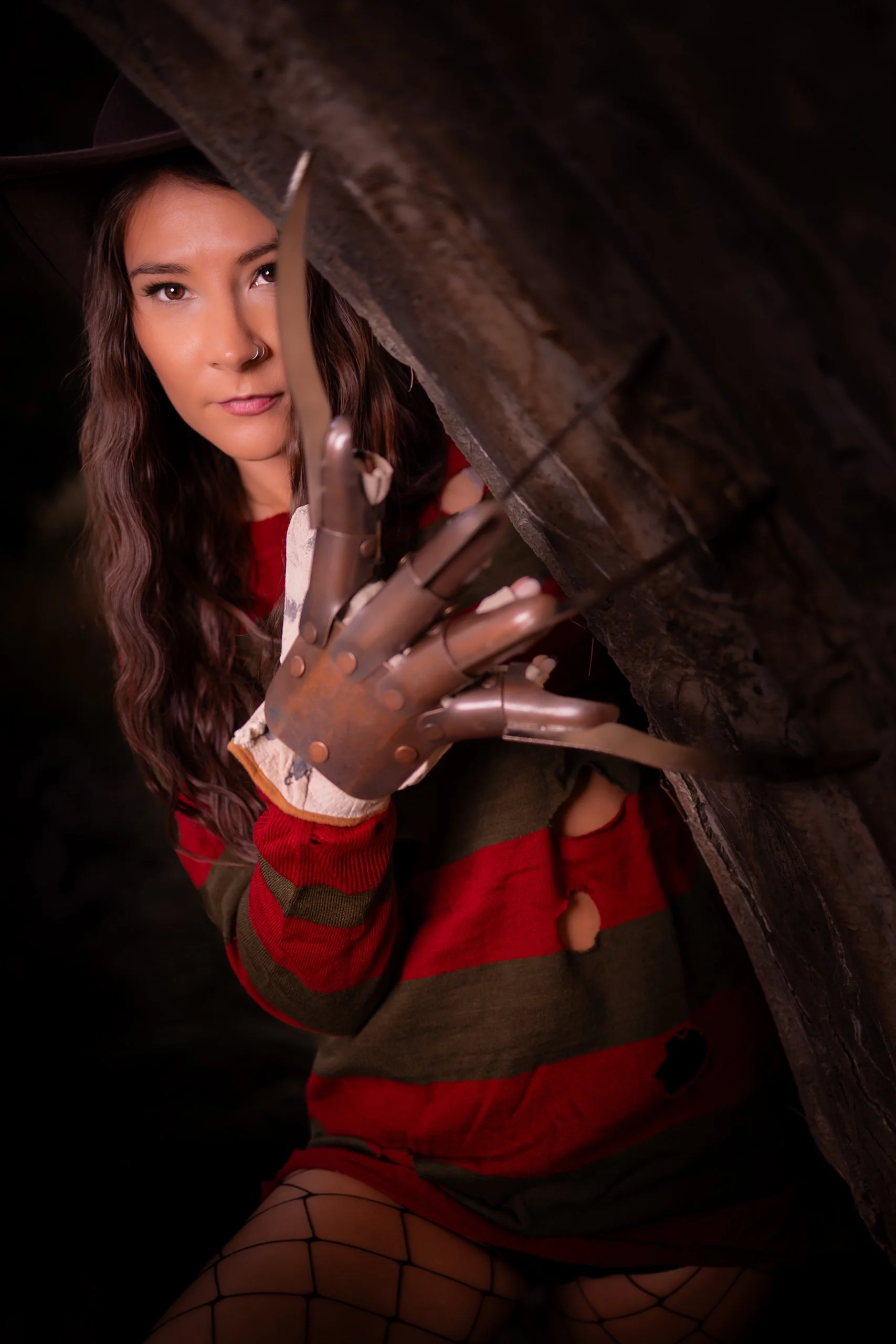 A woman with long wavy hair and a nose ring peeking from behind a rock, wearing a red and green striped sweater, ripped shorts, fishnet stockings, and a robotic glove, looking directly at the camera.