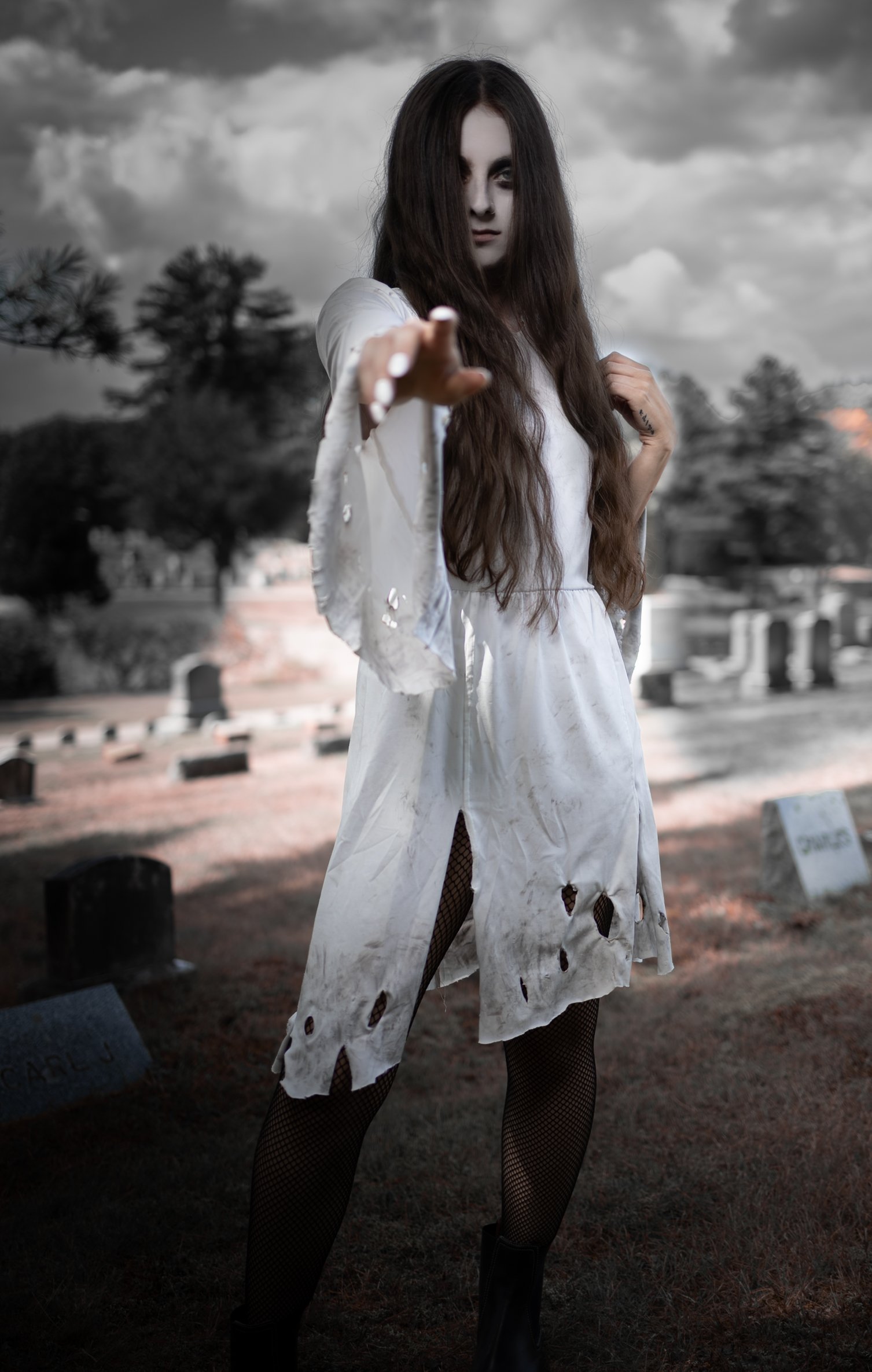 A woman with long hair dressed as a zombie in a dirty white dress with torn fabric, fishnet stockings, and black boots standing in a graveyard, reaching out with her right hand toward the camera.