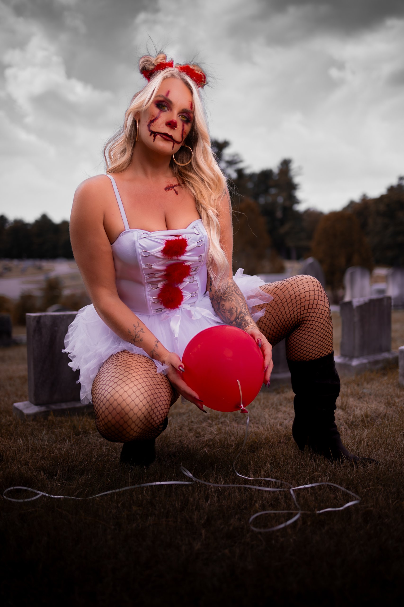 A woman dressed as a creepy clown holding a red balloon in a graveyard during overcast weather.