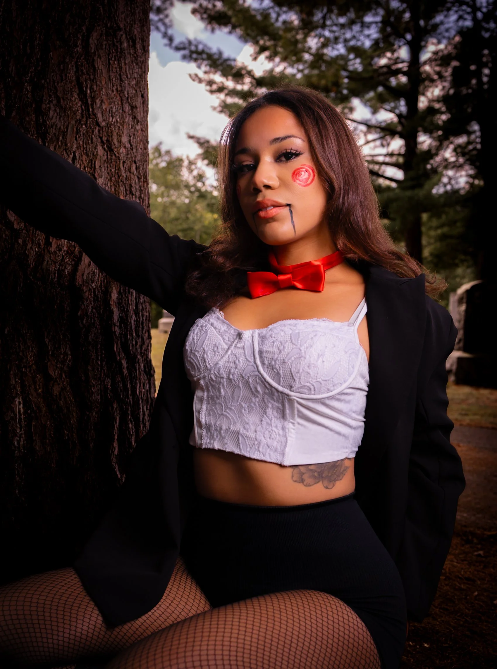 A woman with face paint sits outdoors by a tree, wearing a white lace crop top, black shorts, fishnet stockings, a black blazer, and a red bow tie, with dark hair and makeup.