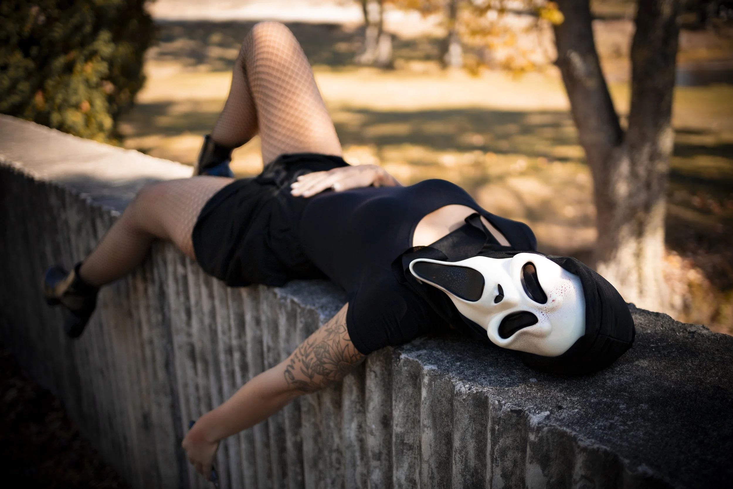 Person lying on a concrete ledge outdoors wearing a white scream mask, black clothing, fishnet stockings, and black shoes, with arms hanging over the edge, in a park-like setting with trees and autumn leaves.