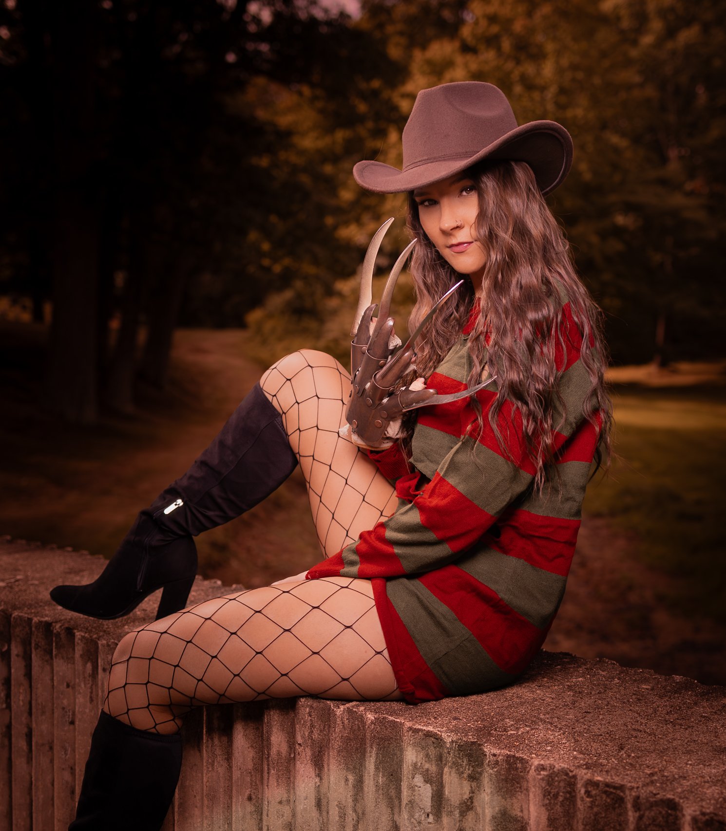 A woman dressed as a horror character, sitting on a brick wall outdoors at sunset, wearing a wide-brimmed hat, striped dress, fishnet stockings, high boots, and a glove with blades, with a forest background.