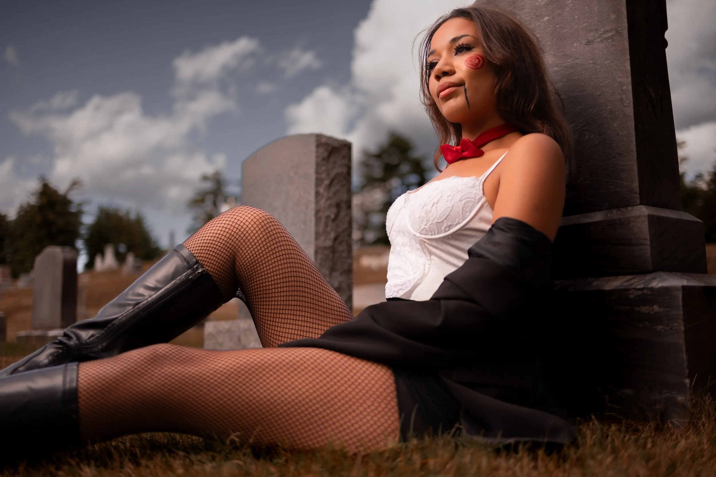 A woman dressed in a costume resembling a Halloween or cosplay outfit, sitting on the ground against a graveyard background with gravestones and trees under a cloudy sky.