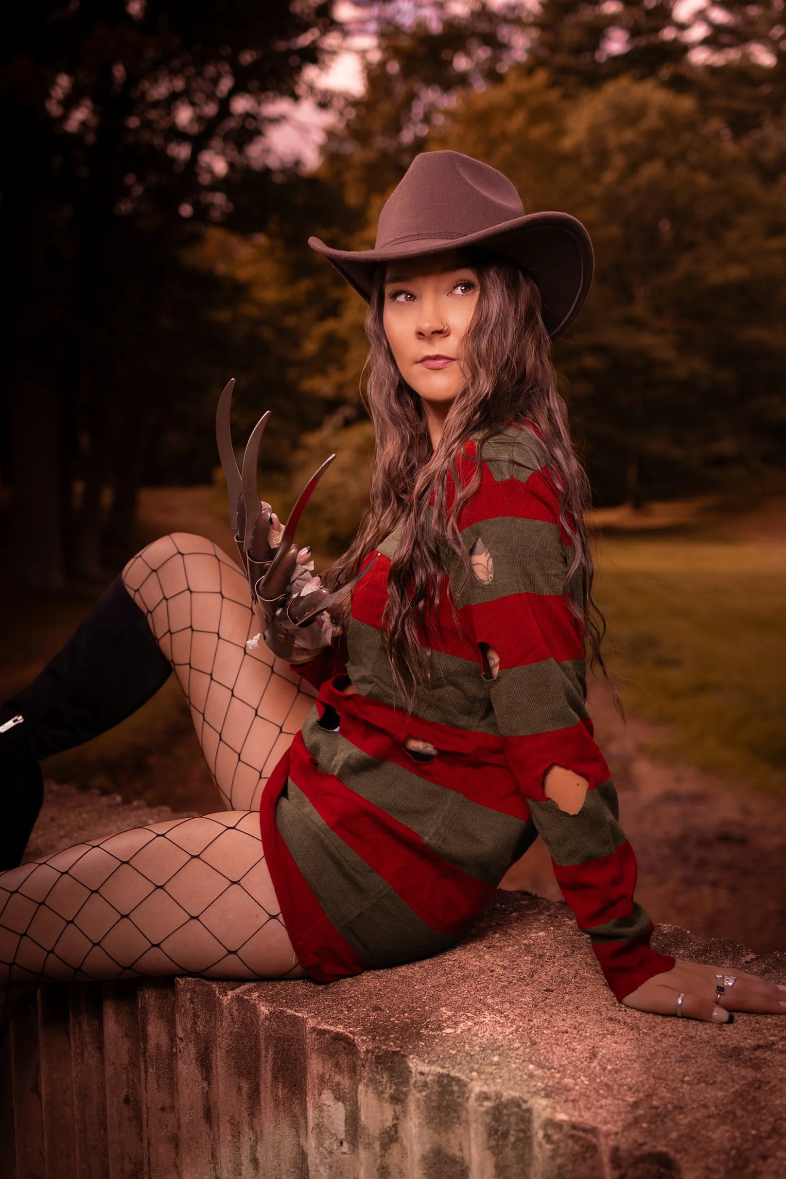 A woman dressed as a witch with a large brown hat, long wavy hair, and costume with ripped red and green striped shirt, fishnet stockings, and holding clawed gloves, sitting outdoors during sunset with trees in the background.
