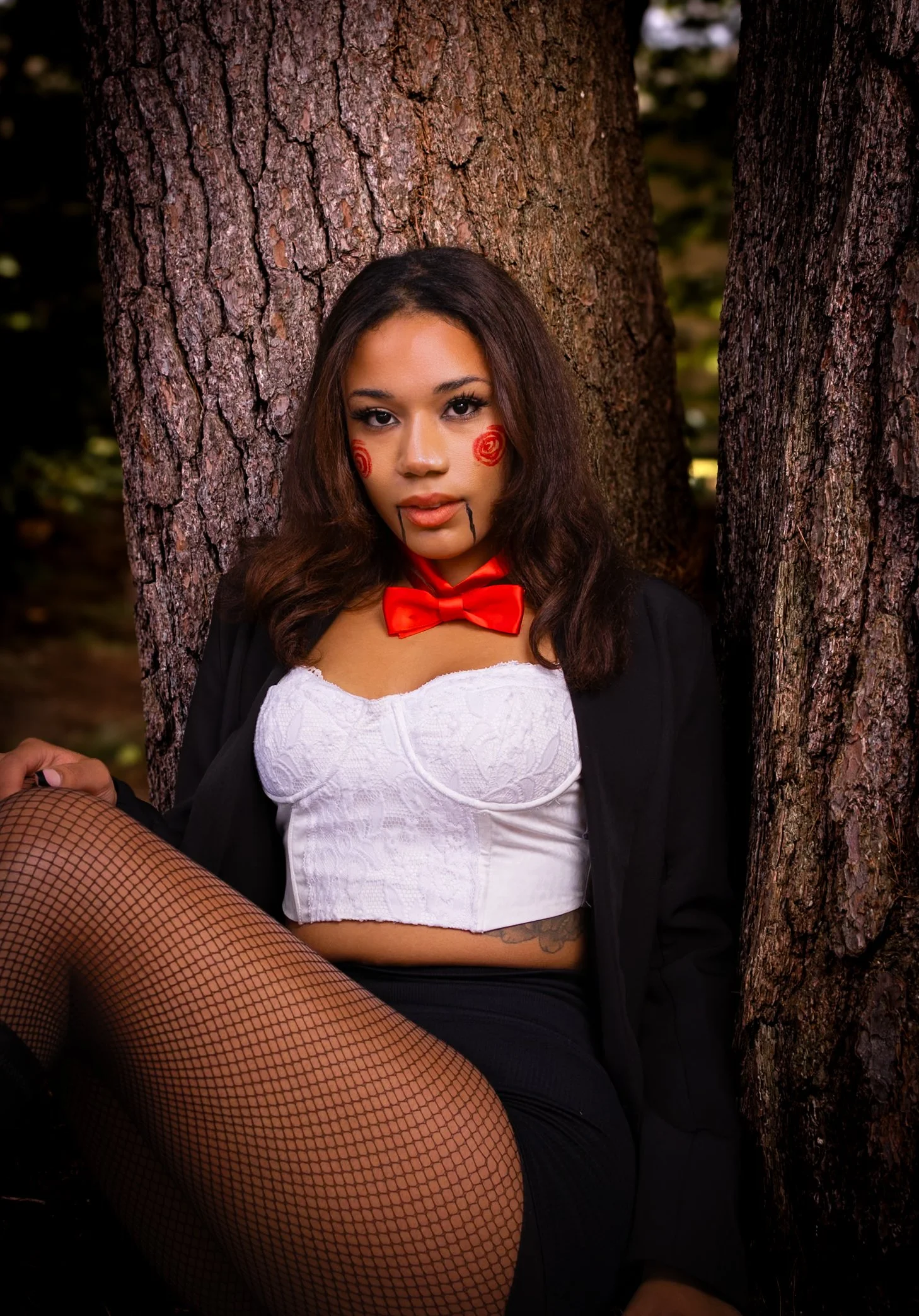 A woman dressed as a clown with face paint, wearing a white lace bustier, black jacket, fishnet stockings, a red bow tie, and red spirals on her cheeks, sitting against a tree in a forest setting.