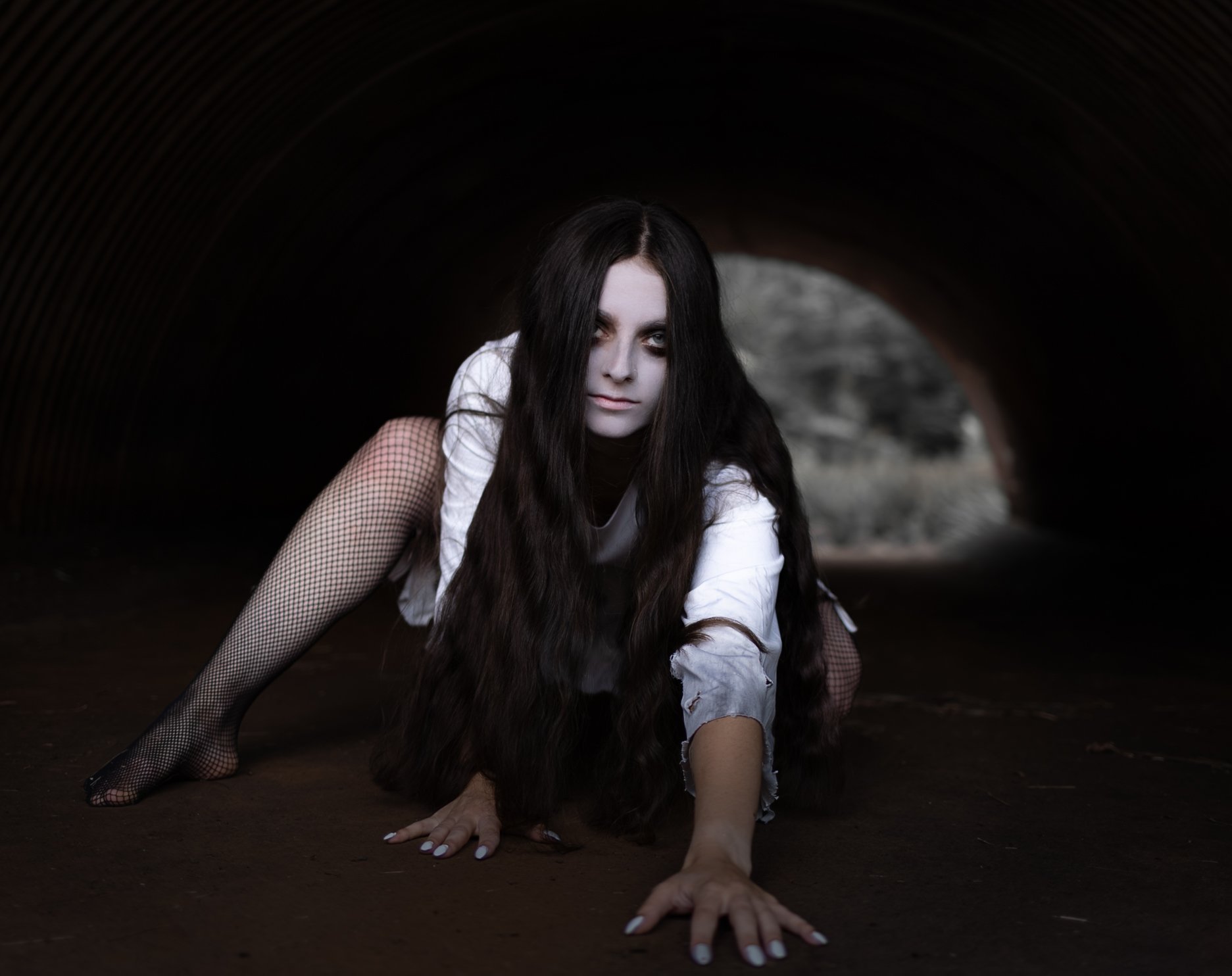 A woman with long dark hair and pale makeup crawling in a dark tunnel, wearing fishnet stockings and distressed white clothing.