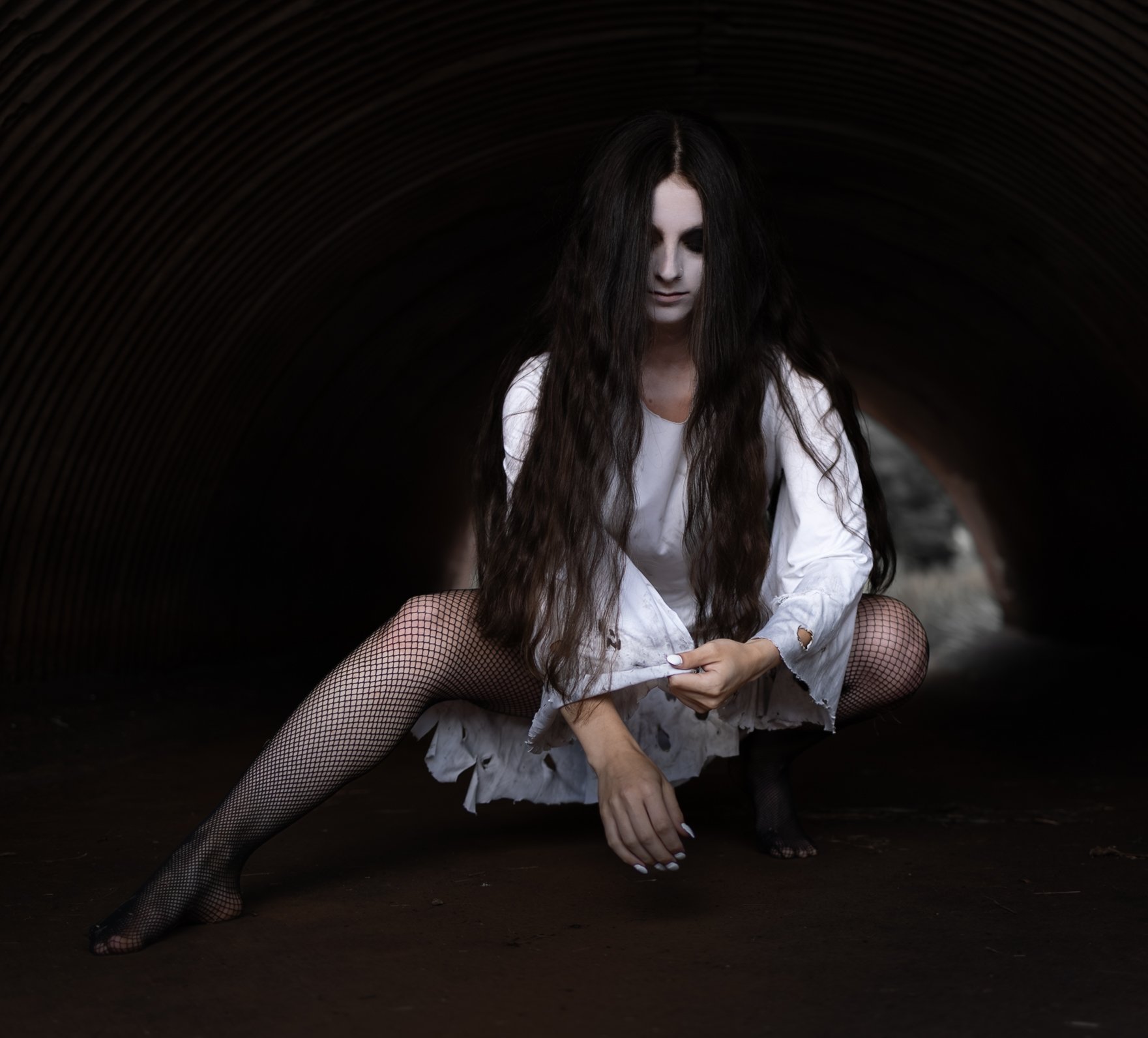 A woman with long dark wavy hair sitting inside a dark tunnel, wearing a white distressed dress and fishnet stockings, in a gothic or edgy style.